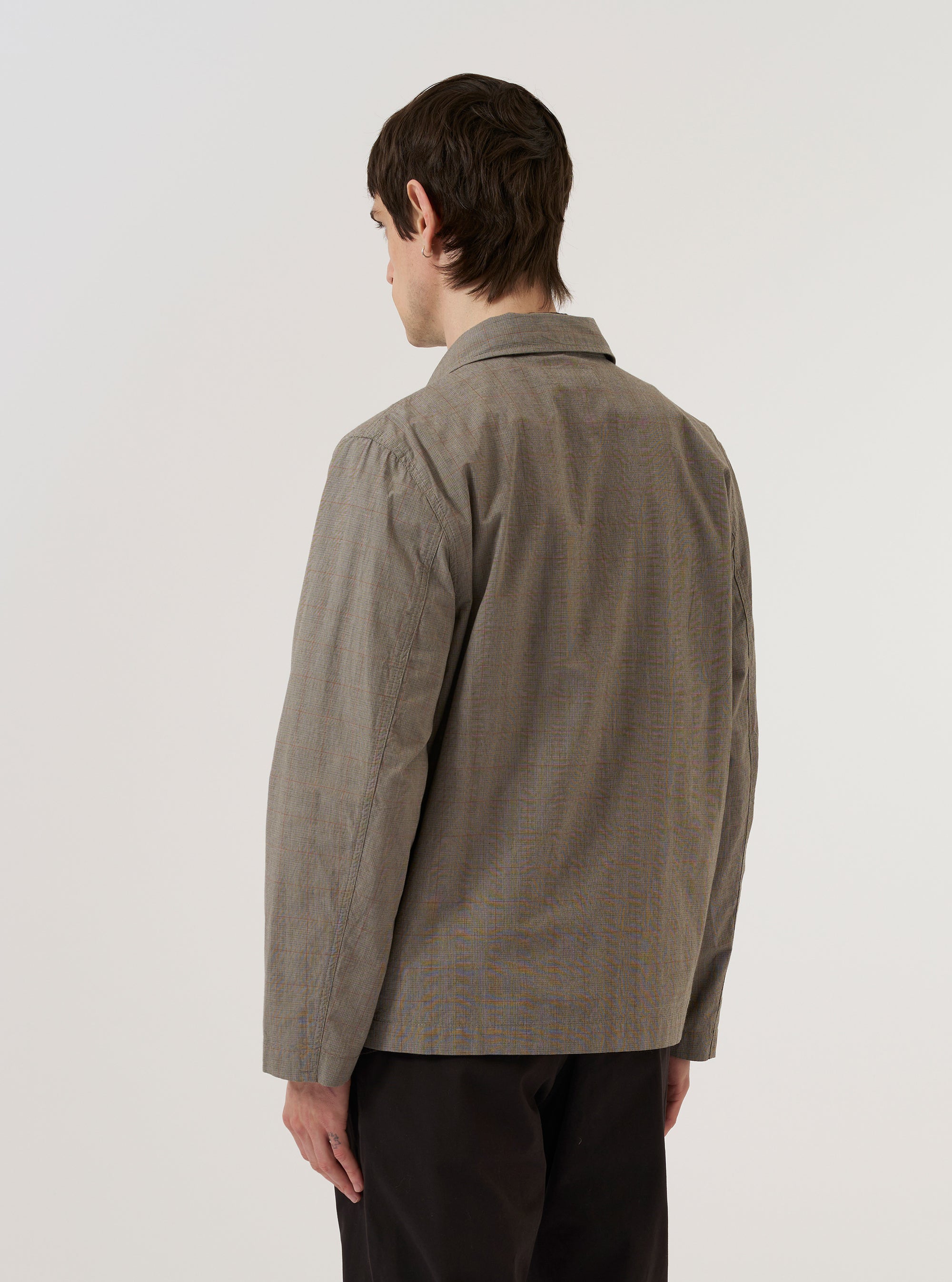 A model wears the Grey Brown Wales Glen Check Franck Jacket, shown from a rear three-quarter angle against a plain, neutral studio background. The jacket features a regular fit, a flat collar, and a straight hem. The intricate grey and brown Glen check pattern is prominent across the cotton fabric.