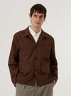 A model is wearing the Brown Peached Poplin Franck Jacket against a white background. The model is shown from the front, and the jacket is worn open over a white shirt. The garment features a flat collar, a single chest welt pocket, and two large patch pockets with flaps at the hips. The jacket has a boxy, regular fit and is constructed from a smooth, brown peached poplin fabric.