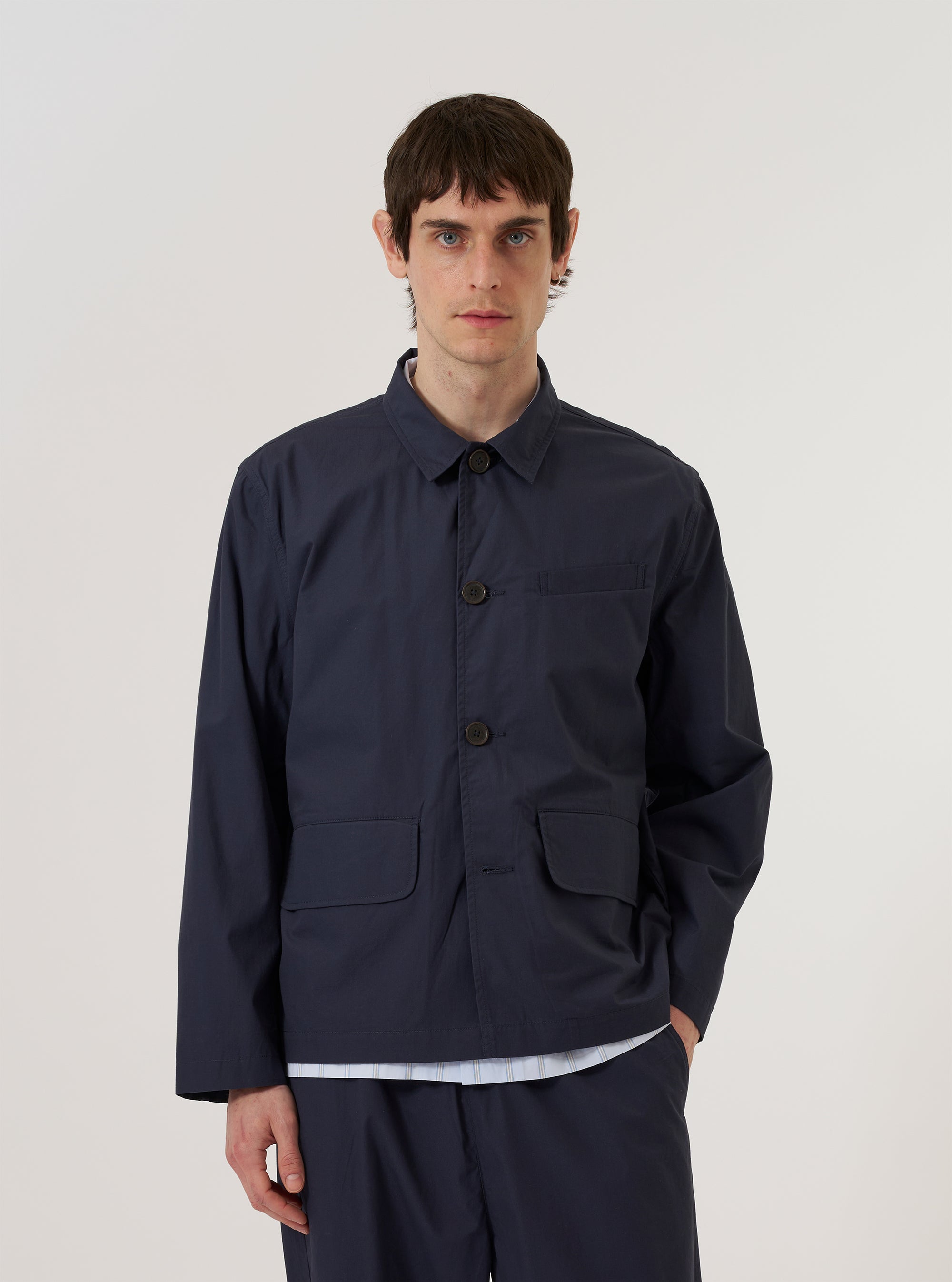 A model wears the Navy Peached Poplin Franck Jacket against a plain white background. The jacket features a one-piece flat collar, a single open chest pocket, and two lower pockets with flap closures. It has a regular fit and is styled open over a light-colored top.