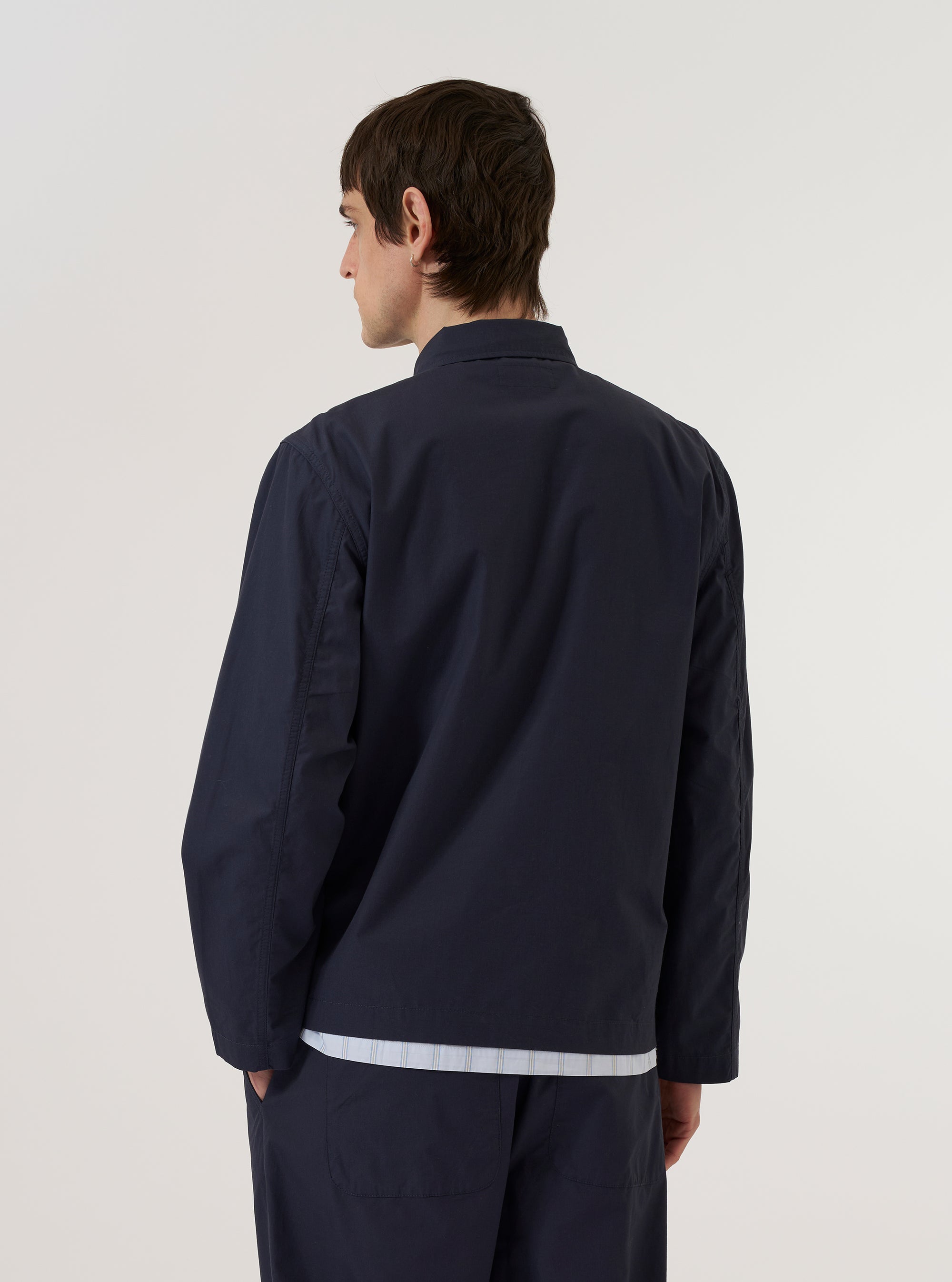 A model wears the Navy Peached Poplin Franck Jacket against a plain, light-colored background. The jacket features a classic collar, a tonal button-down front, and large patch pockets at the waist. The smooth, matte texture of the navy peached poplin fabric is visible. The model is shown from a front-facing perspective to highlight the garment's clean lines and fit.
