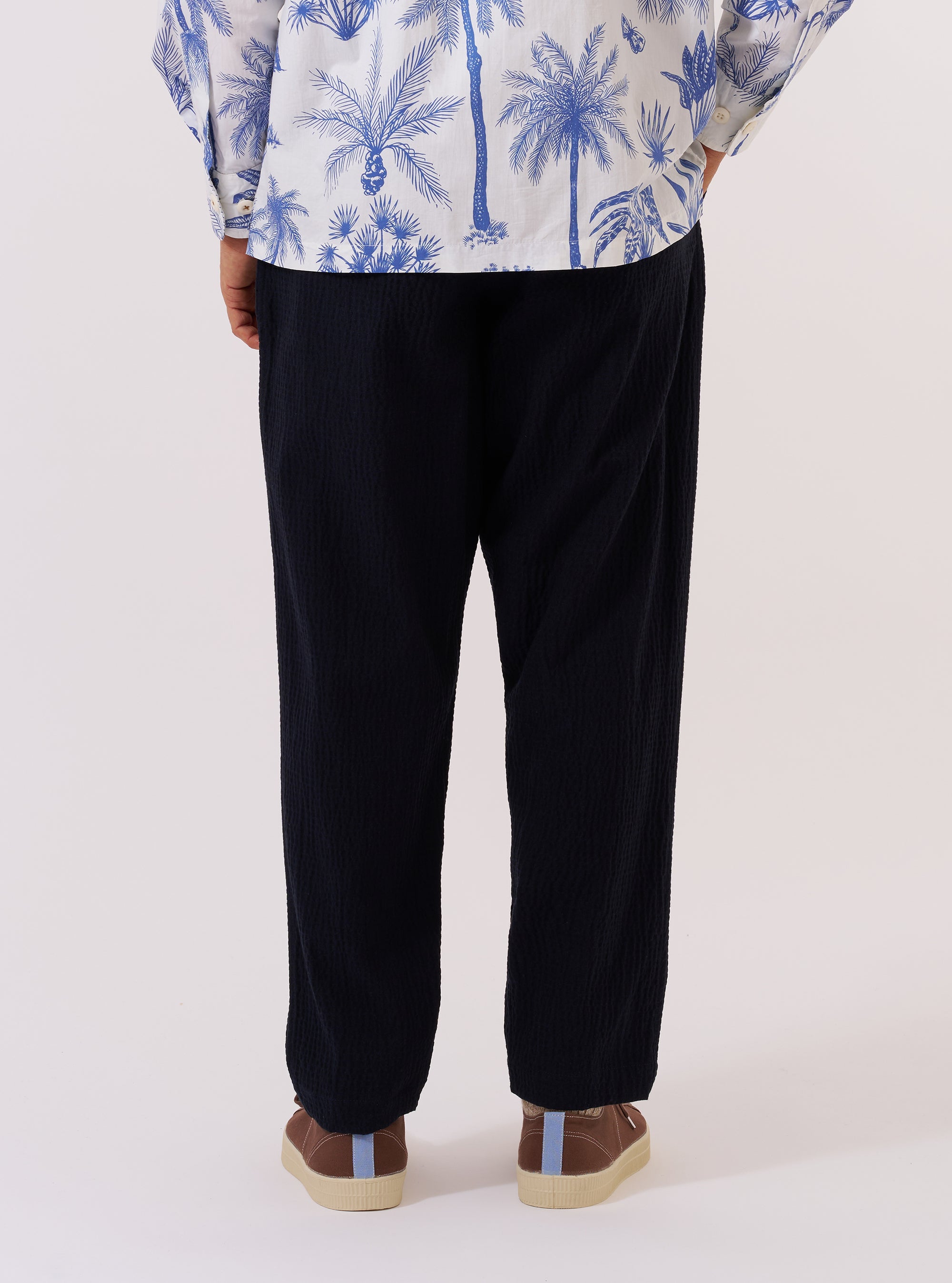 A model is wearing the Dark Navy Ospina Cotton Pleated Track Pant, shown from a rear three-quarter angle against a plain white background. The image highlights the loose, tapered fit of the trousers and the textured dark navy fabric. One rear buttoned flap pocket is visible, along with the elasticated waistband and straight hem. The model is also wearing a light-colored long-sleeve shirt and dark shoes. No mannequin is present in the image.