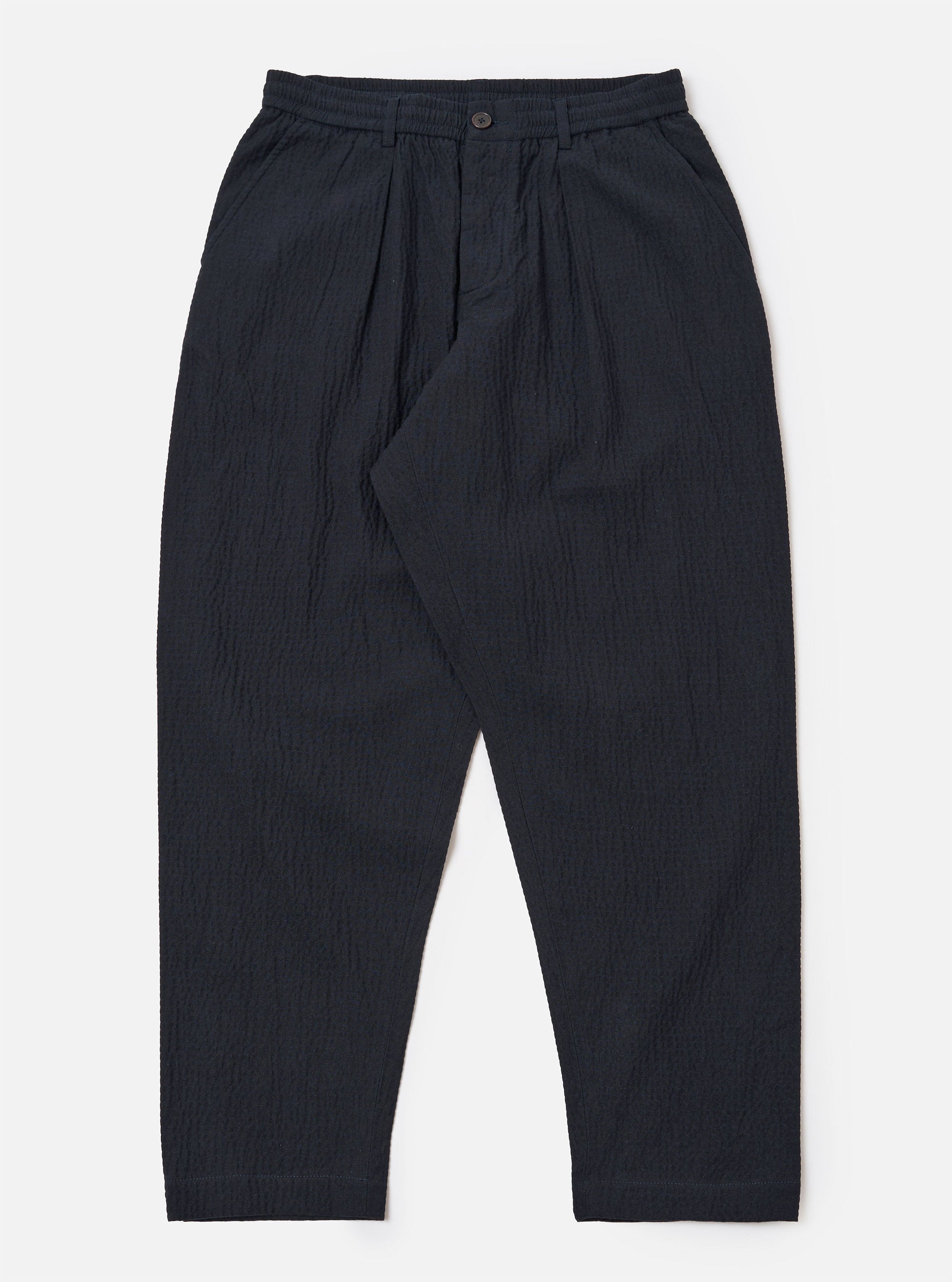 The Dark Navy Ospina Cotton Pleated Track Pant is shown as a flat lay against a plain white background. The trousers feature a single pleat front, an elasticated waistband with a matching drawstring, and a loose fit that tapers toward the hem. The dark navy fabric has a visible seersucker texture.