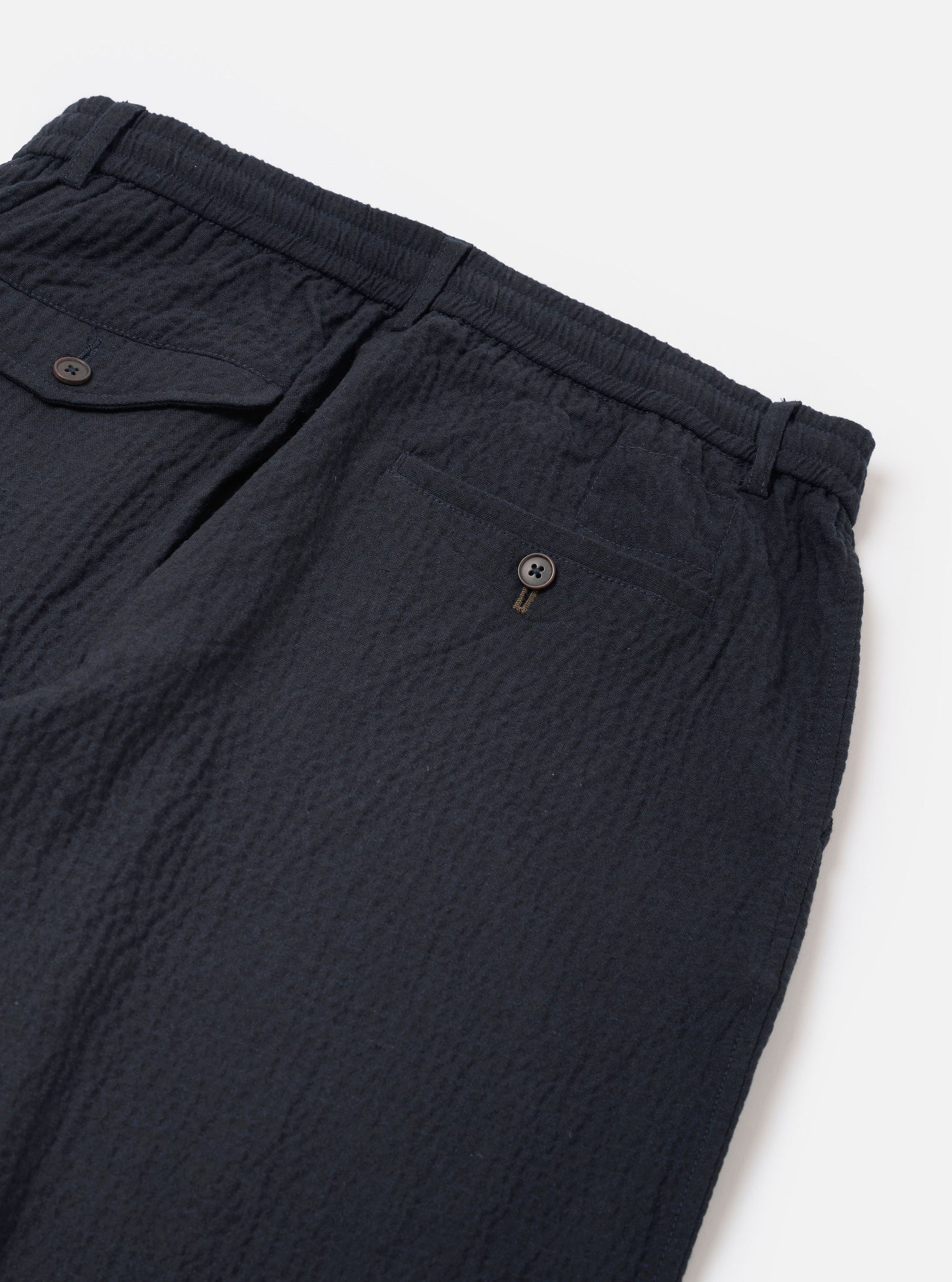 The Dark Navy Ospina Cotton Pleated Track Pant is shown in a detail view as a flat lay against a plain white background. The image highlights the textured seersucker finish of the dark navy fabric and the construction of a functional rear patch pocket with a buttoned flap.