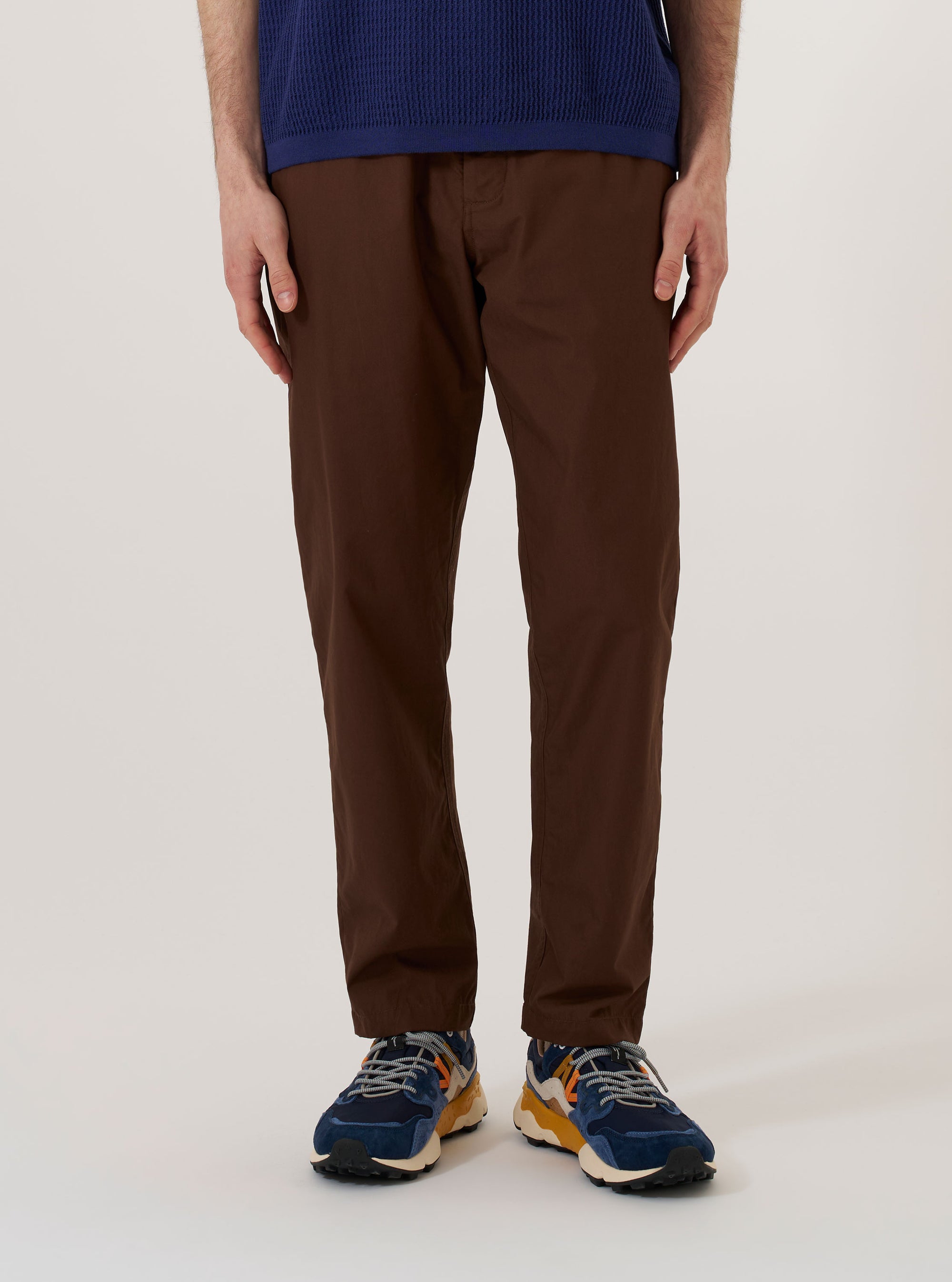 A model wears the Brown Peached Poplin Military Chino while standing against a plain white background. The image shows a front view of the trousers, highlighting the straight-leg fit, side pockets, and the smooth texture of the brown cotton poplin fabric. The model is wearing the chinos with a tucked-in shirt and dark shoes.
