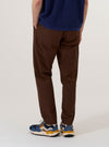 A model wears the Brown Peached Poplin Military Chino, shown from the waist down against a plain, neutral studio background. The trousers feature a slim-straight fit and are constructed from a brown peached poplin fabric with a soft, matte finish. This rear three-quarter view highlights the garment's construction, including the waistband with belt loops and two button-through rear pockets. The model is wearing a light-colored top tucked into the chinos.