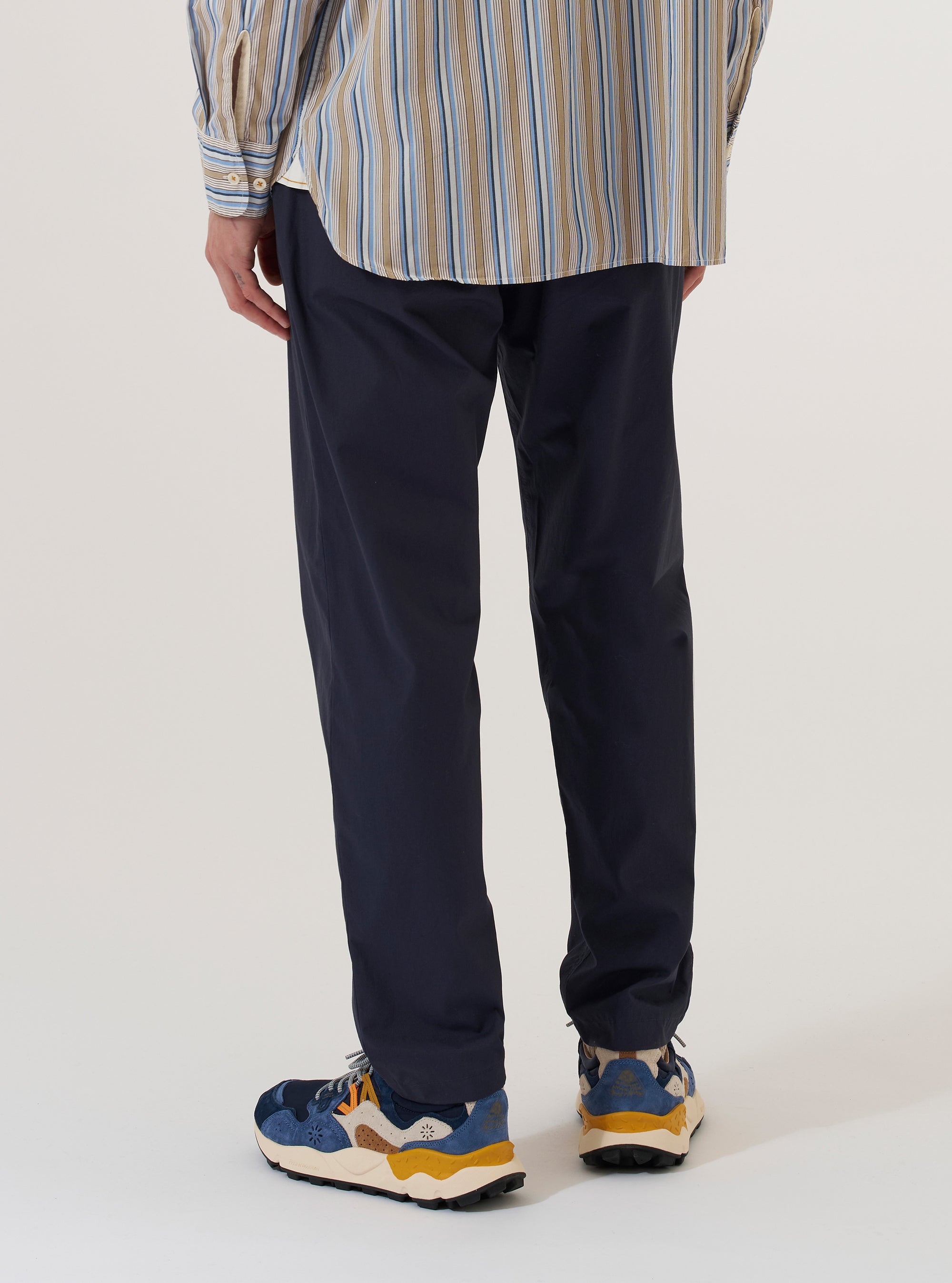 A model is wearing the Navy Peached Poplin Military Chino, shown from the waist down from a rear angle against a plain white background. The trousers feature a straight-leg silhouette and a rear flap pocket with a button closure. The navy blue fabric has a smooth, matte peached poplin finish.