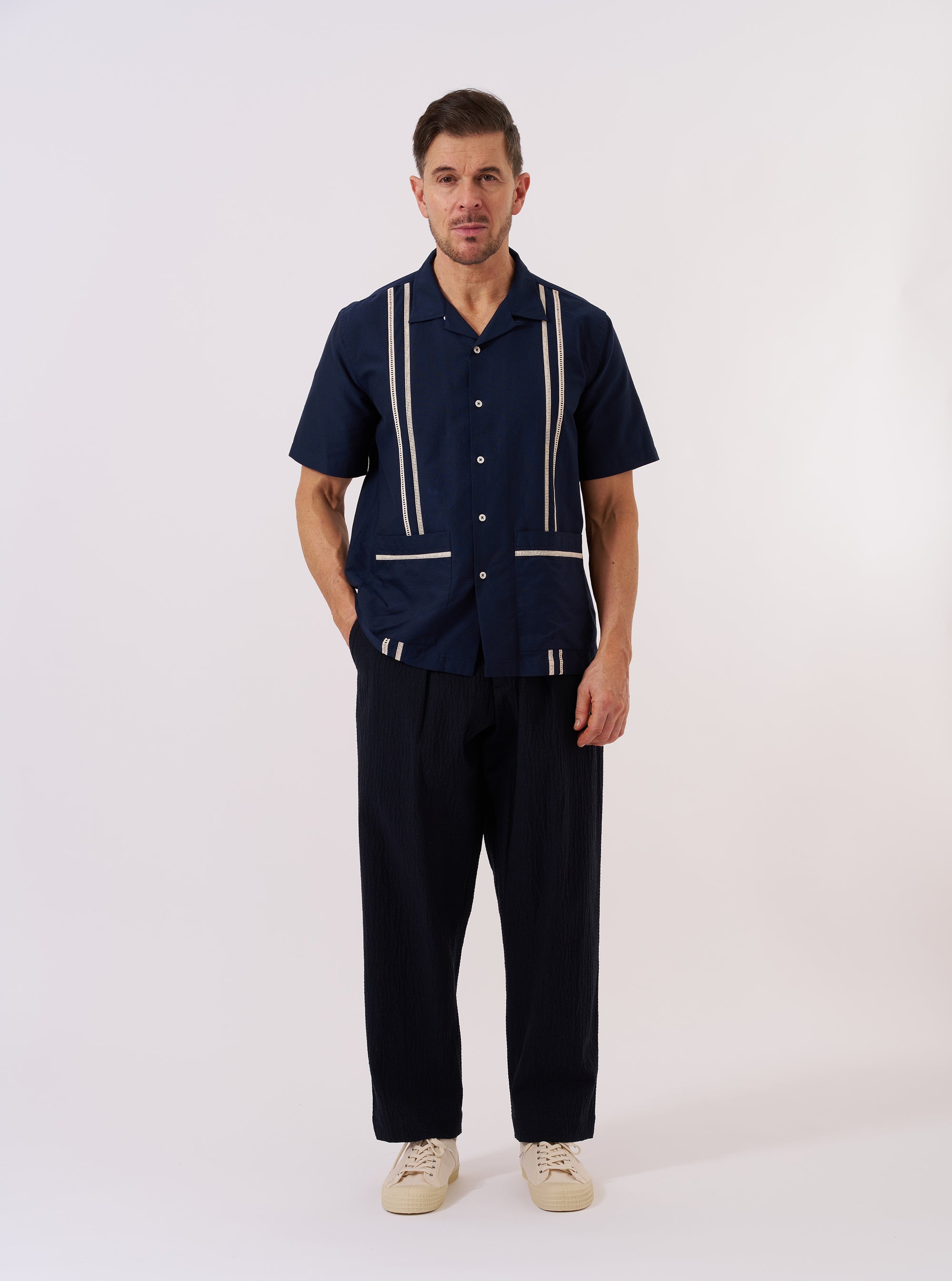 The Navy Classic Oxford Tropicana Shirt is displayed as a flat lay against a plain white background. The shirt features a one-piece open collar, a full-button front, and short sleeves. Two large patch pockets are positioned at the hip, and the hem includes side splits. The navy cotton fabric has a visible oxford weave.
