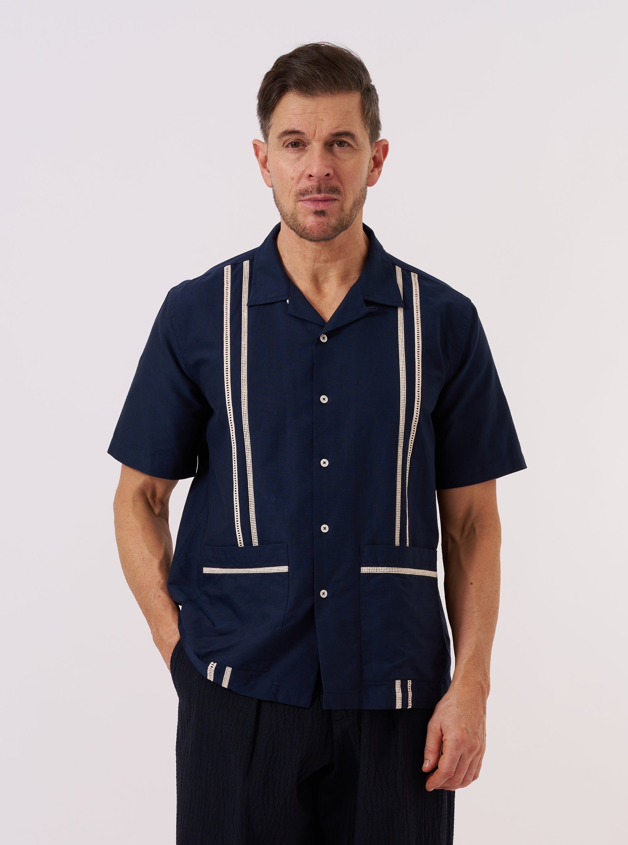 A model wears the Navy Classic Oxford Tropicana Shirt over a light-colored top, standing against a plain white background. The shirt features an open collar, a central button-down closure, and two large patch pockets at the waist. The navy blue cotton Oxford fabric is shown with a relaxed fit and wide short sleeves.
