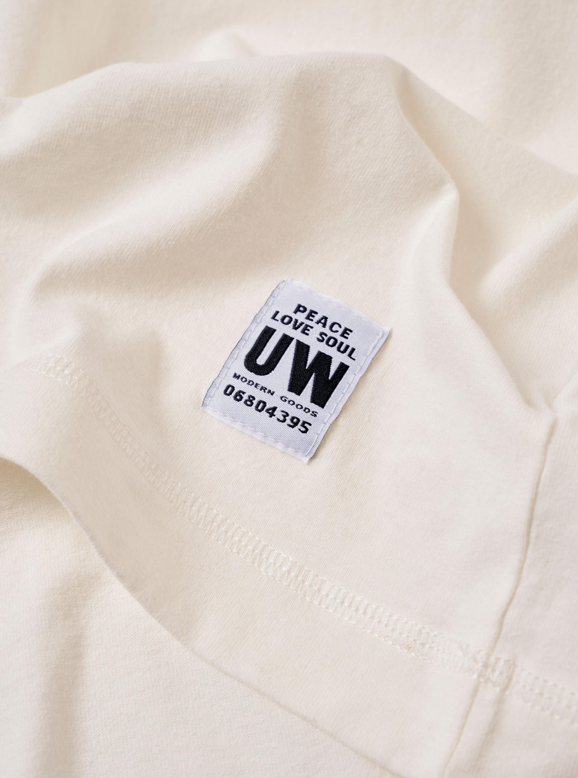 The Antique White Recycled/Bio Jersey Henry L/S Tee is shown from a rear angle as a flat lay against a plain white background. The image highlights the simple, clean construction of the back of the garment, featuring a straight hem and long sleeves. The fabric is a solid antique white jersey.