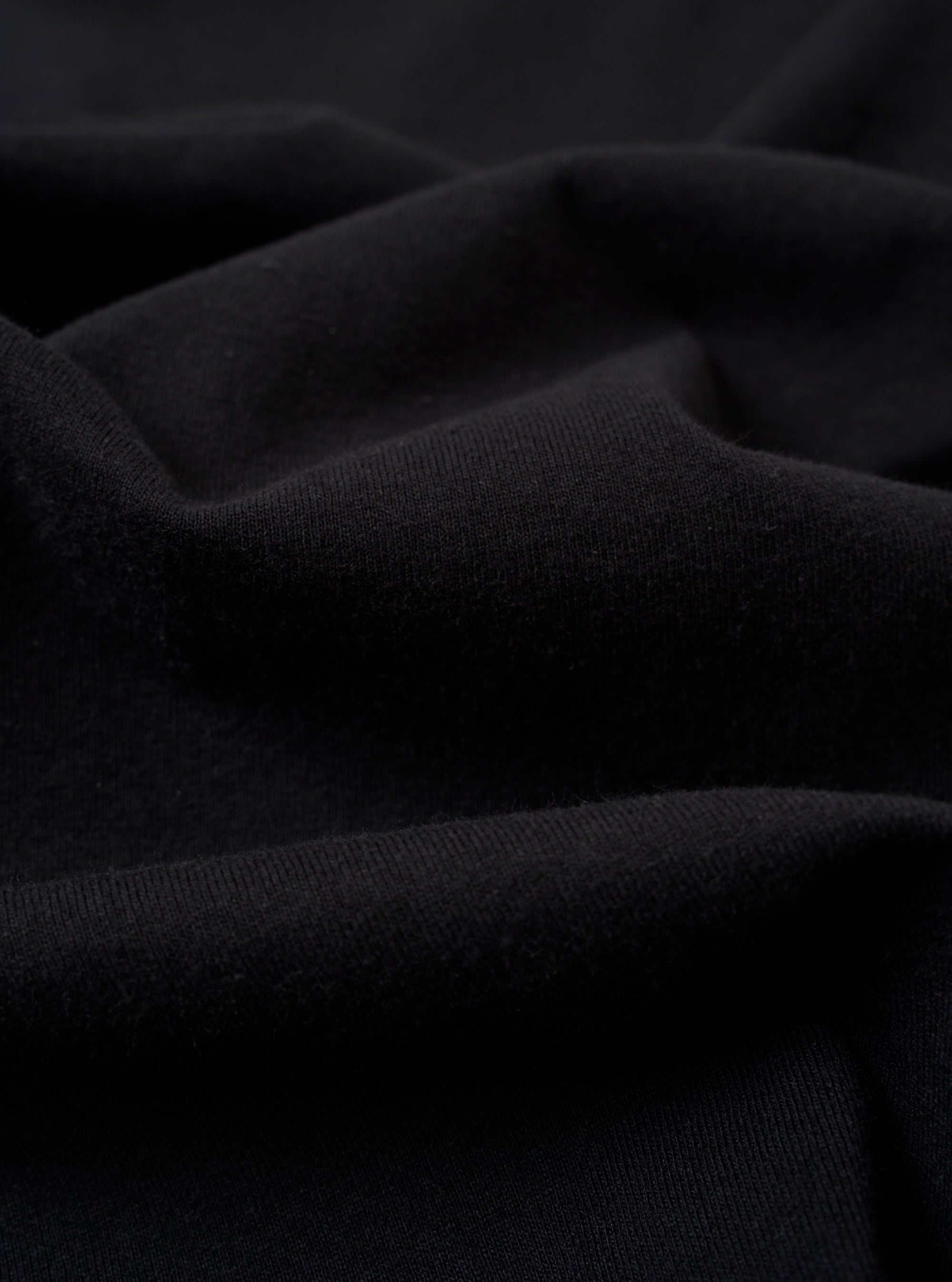 A close-up fabric swatch of the Black Recycled/Bio Jersey Henry L/S Tee displays the solid black color and the fine, soft knit texture of the recycled and organic cotton jersey material. The fabric fills the entire frame.