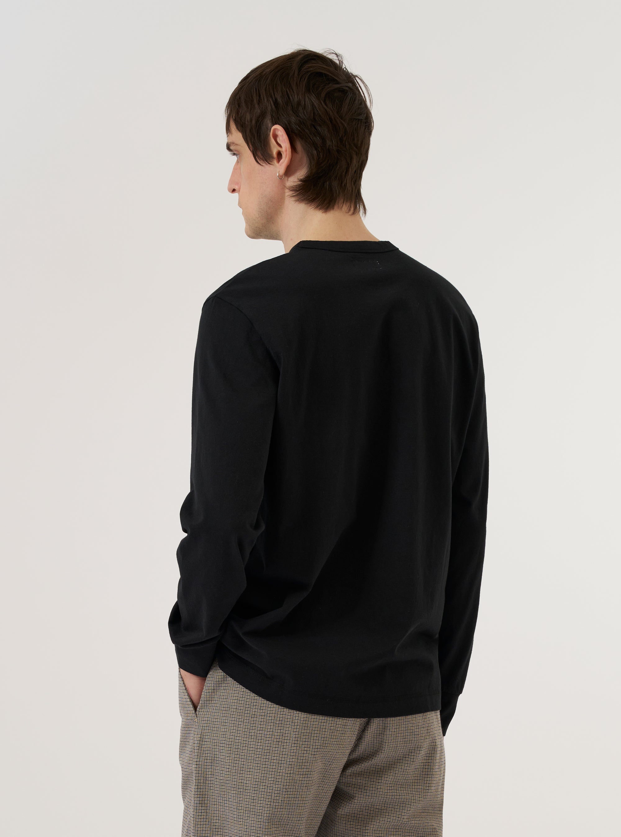 A model wears the Black Recycled/Bio Jersey Henry L/S Tee against a plain, neutral background. The long-sleeve T-shirt features a classic crew neckline and a relaxed fit, highlighting the smooth, matte texture of the black organic and recycled cotton jersey fabric. The garment is shown in a studio setting, emphasizing its clean lines and simple construction.