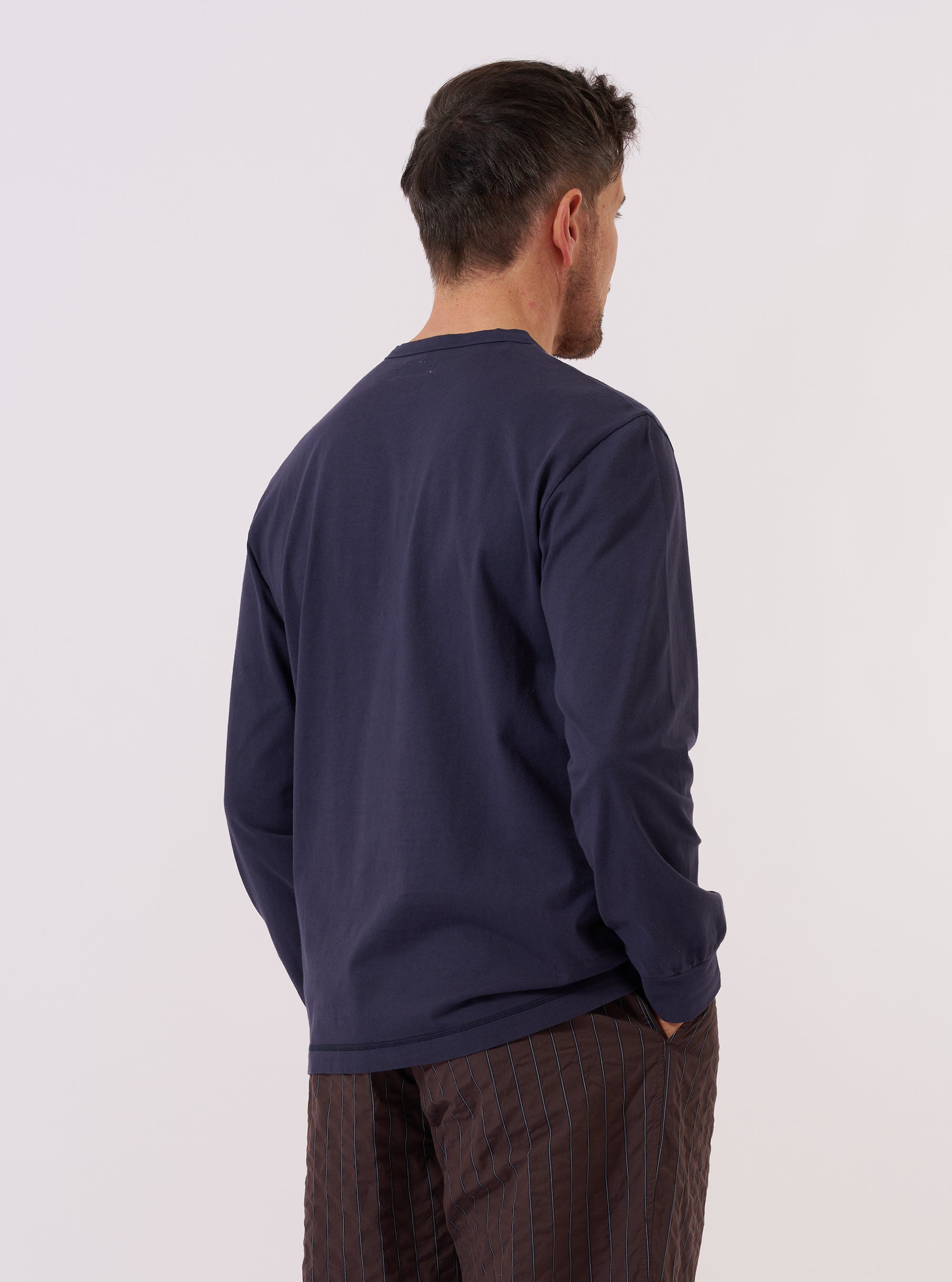 A model wears the Navy Recycled/Bio Jersey Henry L/S Tee, shown from the back against a plain white background. The long-sleeved garment features a relaxed fit and a straight hem, highlighting the soft texture and deep blue color of the recycled and organic cotton jersey fabric. No other accessories are prominent in this rear-view studio shot.