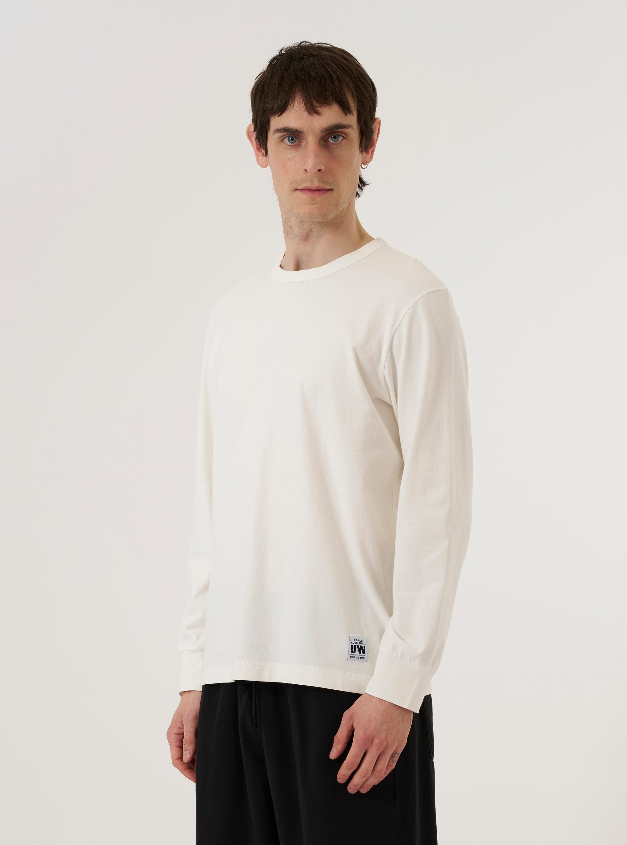 A model wears the Antique White Recycled/Bio Jersey Henry L/S Tee against a plain white background. The long-sleeved top features a three-button front placket at the neckline and is made from a smooth, off-white jersey fabric. The model is shown from the front, highlighting the garment's relaxed fit and clean construction.