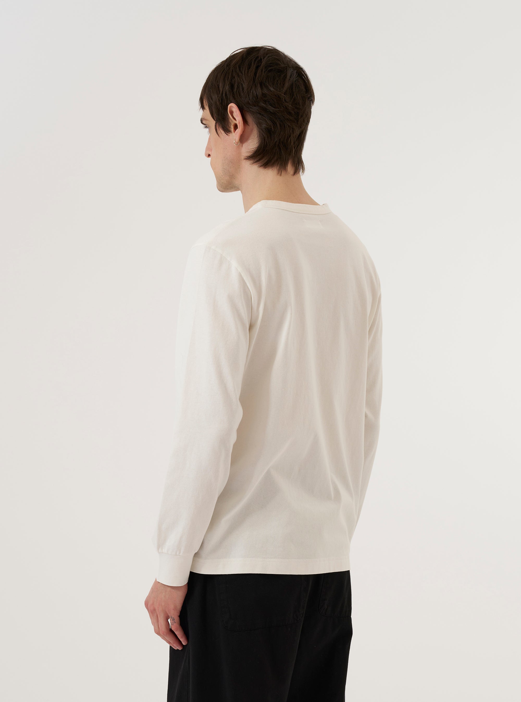 A model wears the Antique White Recycled/Bio Jersey Henry L/S Tee, shown from a rear three-quarter angle against a plain, neutral background. The description highlights the long sleeves, the straight hem, and the soft, matte texture of the antique white jersey fabric. The garment features a relaxed fit with clean stitching details visible along the seams.