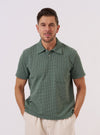 A model wears the Green Argyll Jersey Newlyn Polo against a plain, light-colored background. The polo shirt features a prominent green and navy blue argyll diamond pattern. It is designed with a classic flat collar, a short button placket, and ribbed cuffs on the short sleeves. The model is shown from the waist up, demonstrating the fit and the textured jersey knit of the fabric.
