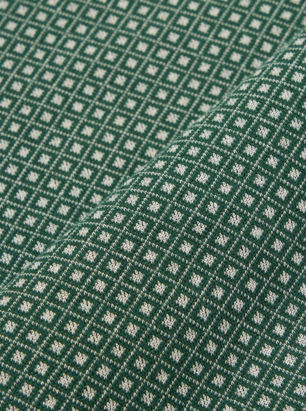 A close-up fabric swatch of the Green Argyll Jersey Newlyn Polo shows a green textile with a subtle, textured argyll pattern. The fabric is displayed flat, highlighting the intricate knit detail and varying shades of green.