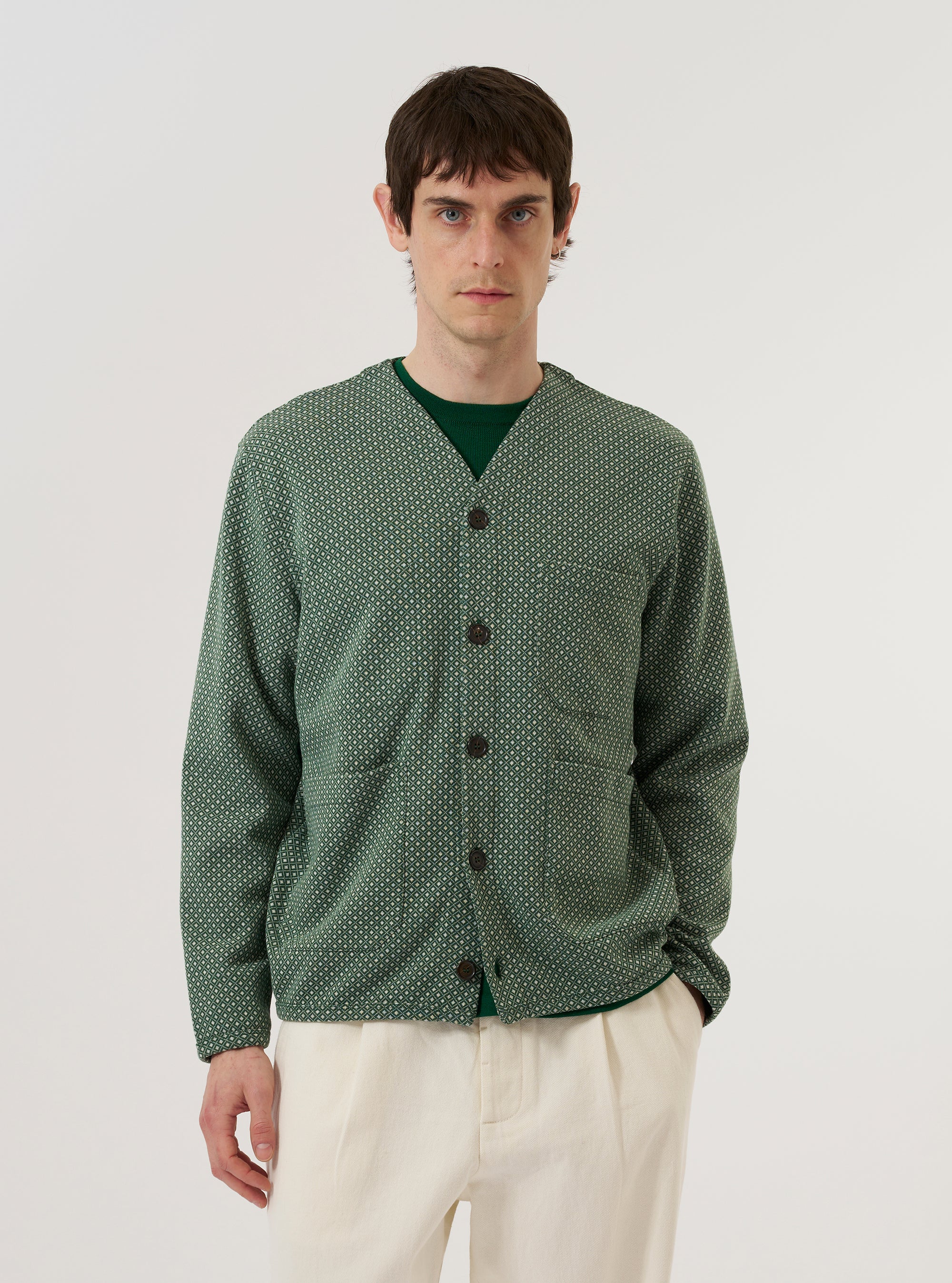 A model wears the Green Argyll Jersey Cardigan against a plain white background. The cardigan features an all-over tonal green Argyll diamond pattern, a five-button front closure, and ribbed detailing at the V-neckline, cuffs, and hem. The model is shown from the front, wearing the garment over a light-colored t-shirt.