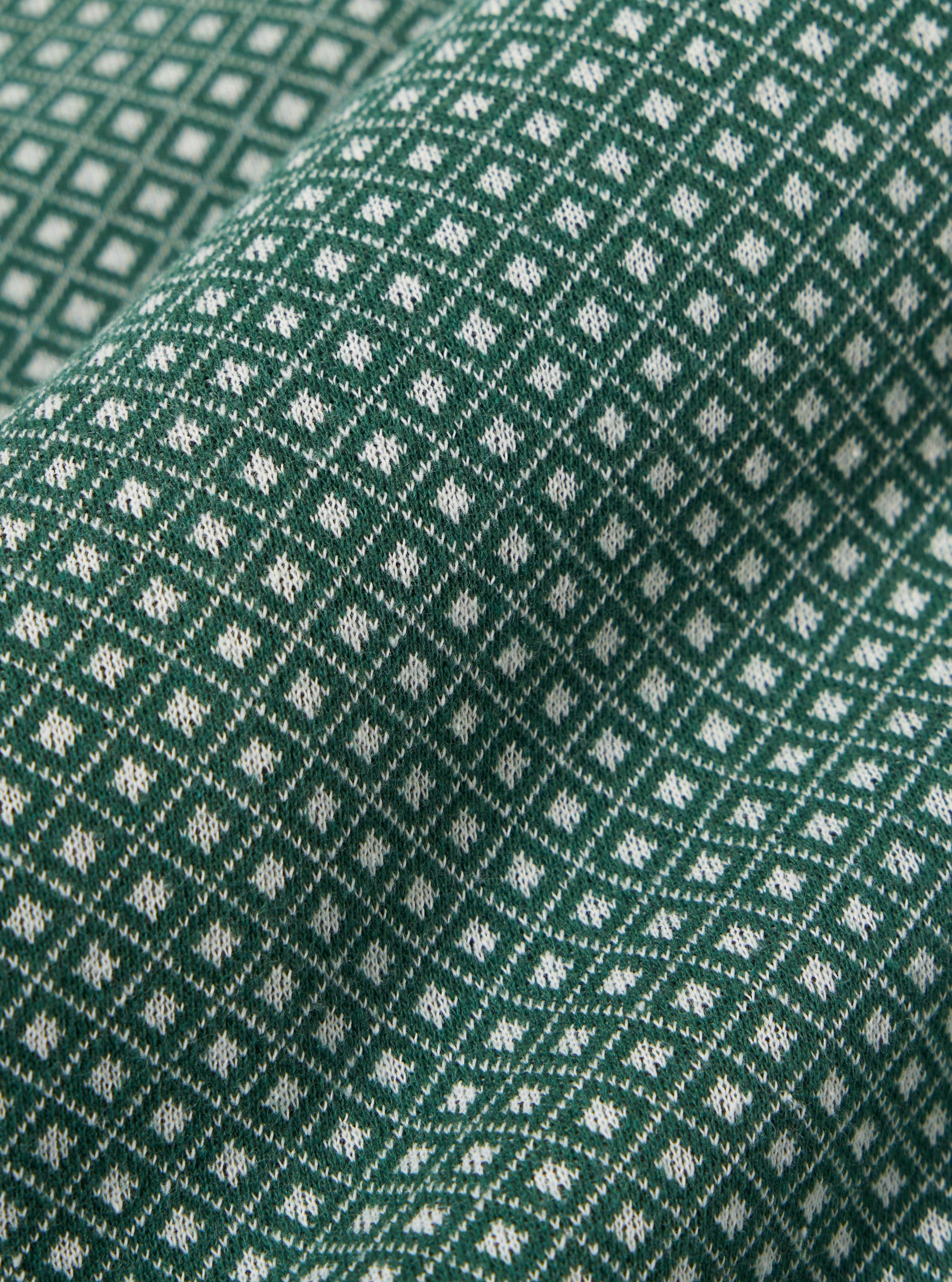 A close-up fabric swatch of the Green Argyll Jersey Cardigan highlights the intricate argyle pattern featuring diamond shapes in shades of green and olive. The image focuses on the soft, knitted texture of the jersey fabric.