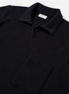 The Black Fantasy Checkerboard Vacation Polo is shown in a close-up detail shot as a flat lay against a plain white background. The image focuses on the upper portion of the garment, highlighting the open-neck design with a short placket, a single-piece collar, and a single patch chest pocket. The organic cotton fabric features a prominent black and grey fantasy checkerboard pattern.