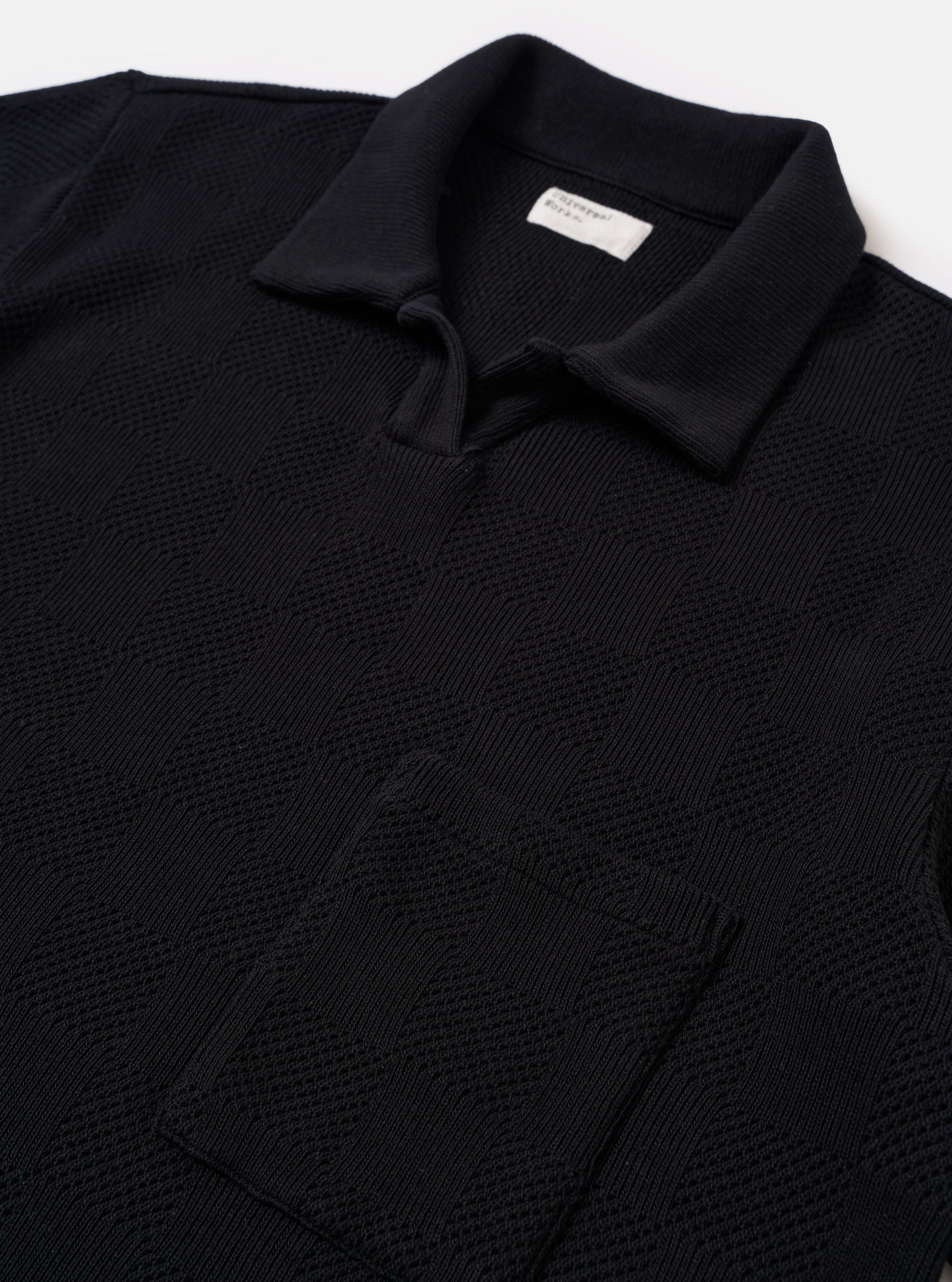The Black Fantasy Checkerboard Vacation Polo is shown in a close-up detail shot as a flat lay against a plain white background. The image focuses on the upper portion of the garment, highlighting the open-neck design with a short placket, a single-piece collar, and a single patch chest pocket. The organic cotton fabric features a prominent black and grey fantasy checkerboard pattern.
