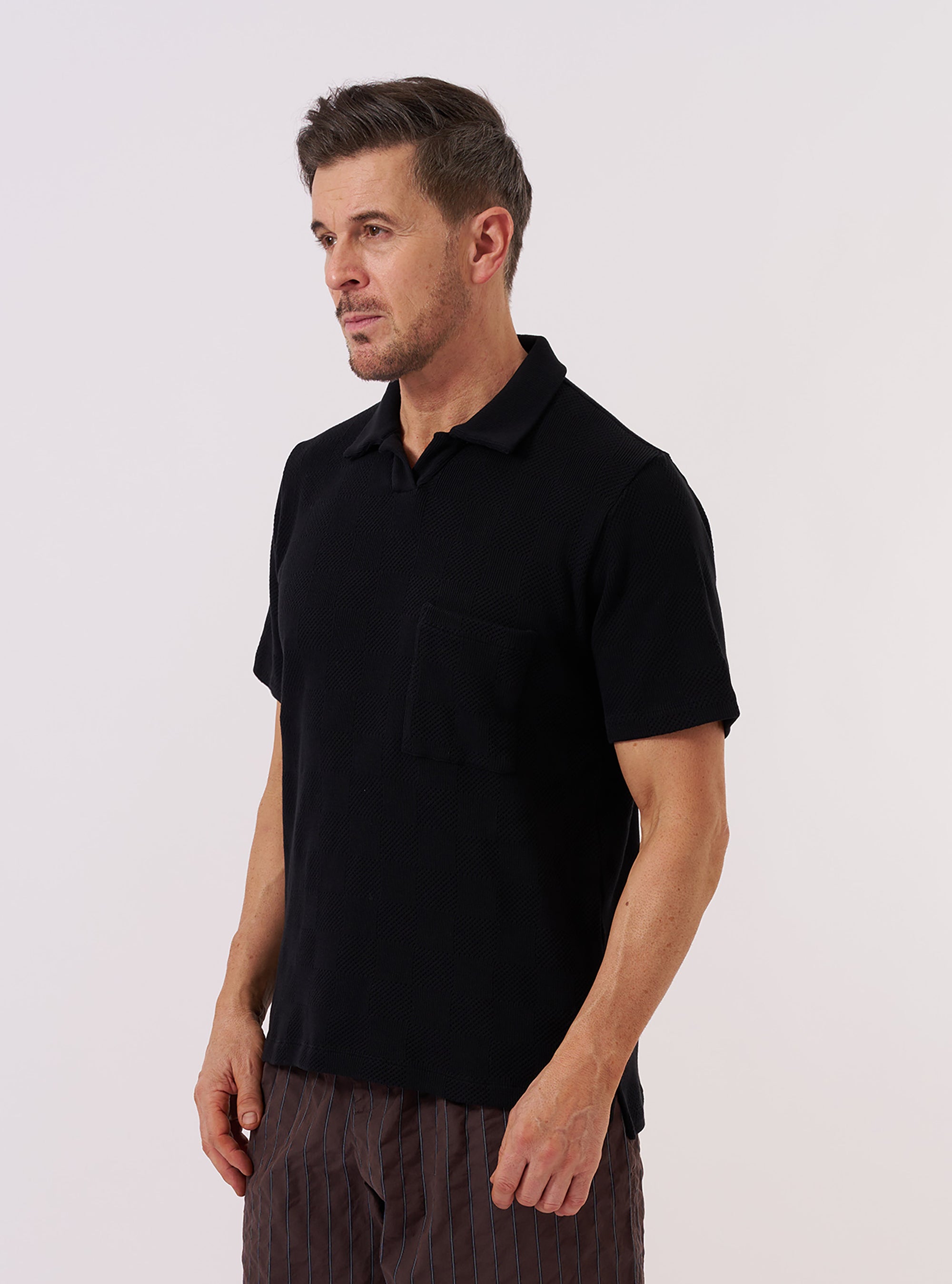 A model wears the Black Fantasy Checkerboard Vacation Polo against a plain white background. The polo features an all-over black and grey checkerboard pattern, a relaxed open neck with a one-piece collar, and short sleeves. The model is shown from the chest up, highlighting the garment's casual silhouette and intricate knit texture.