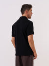 A model wears the Black Fantasy Checkerboard Vacation Polo, shown from a side angle against a plain, neutral studio background. The polo features an all-over black and grey checkerboard pattern, a relaxed fit, and a classic vacation collar. The textured organic cotton fabric and the short sleeves are prominent features in the image.