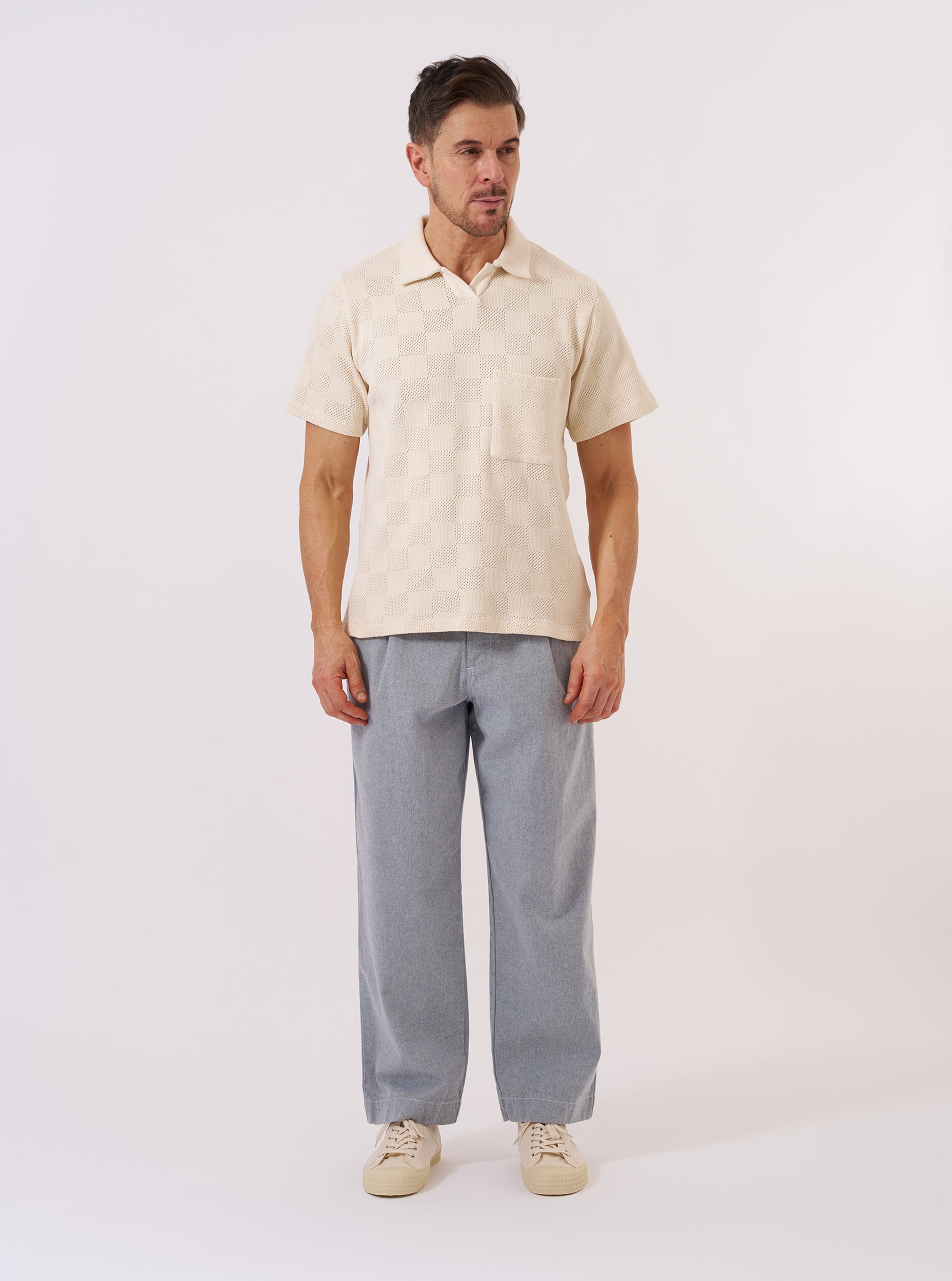 The Ecru Fantasy Checkerboard Vacation Polo is shown as a flat lay against a plain white background. The polo features a regular fit, a classic collar, a three-button placket, and a short-sleeved design with a split hem. The organic cotton fabric is decorated with an all-over fantasy checkerboard pattern in ecru and white.
