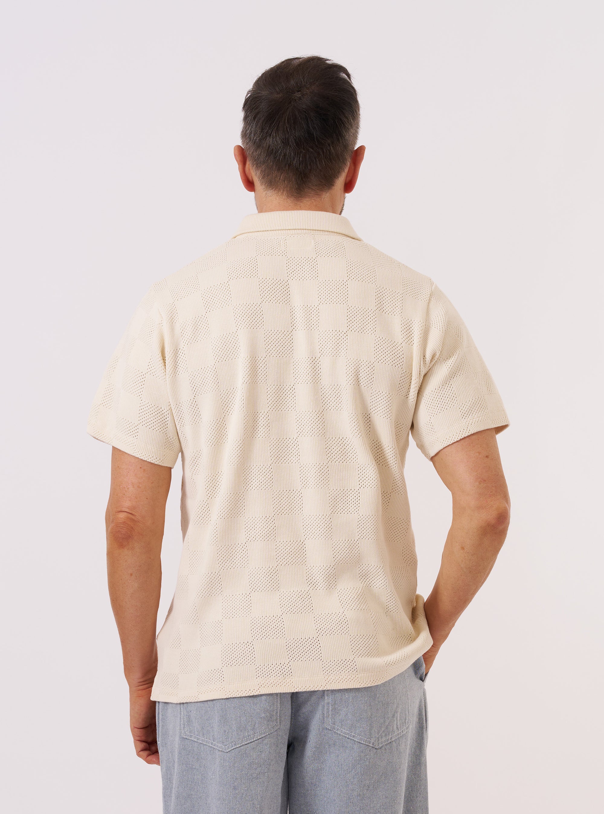 A model is wearing the Ecru Fantasy Checkerboard Vacation Polo, shown from the back against a plain white background. The polo features a relaxed fit, a classic collar, and short sleeves, showcasing the intricate ecru and white checkerboard knit pattern across the entire back. The model is standing upright with their back to the camera.