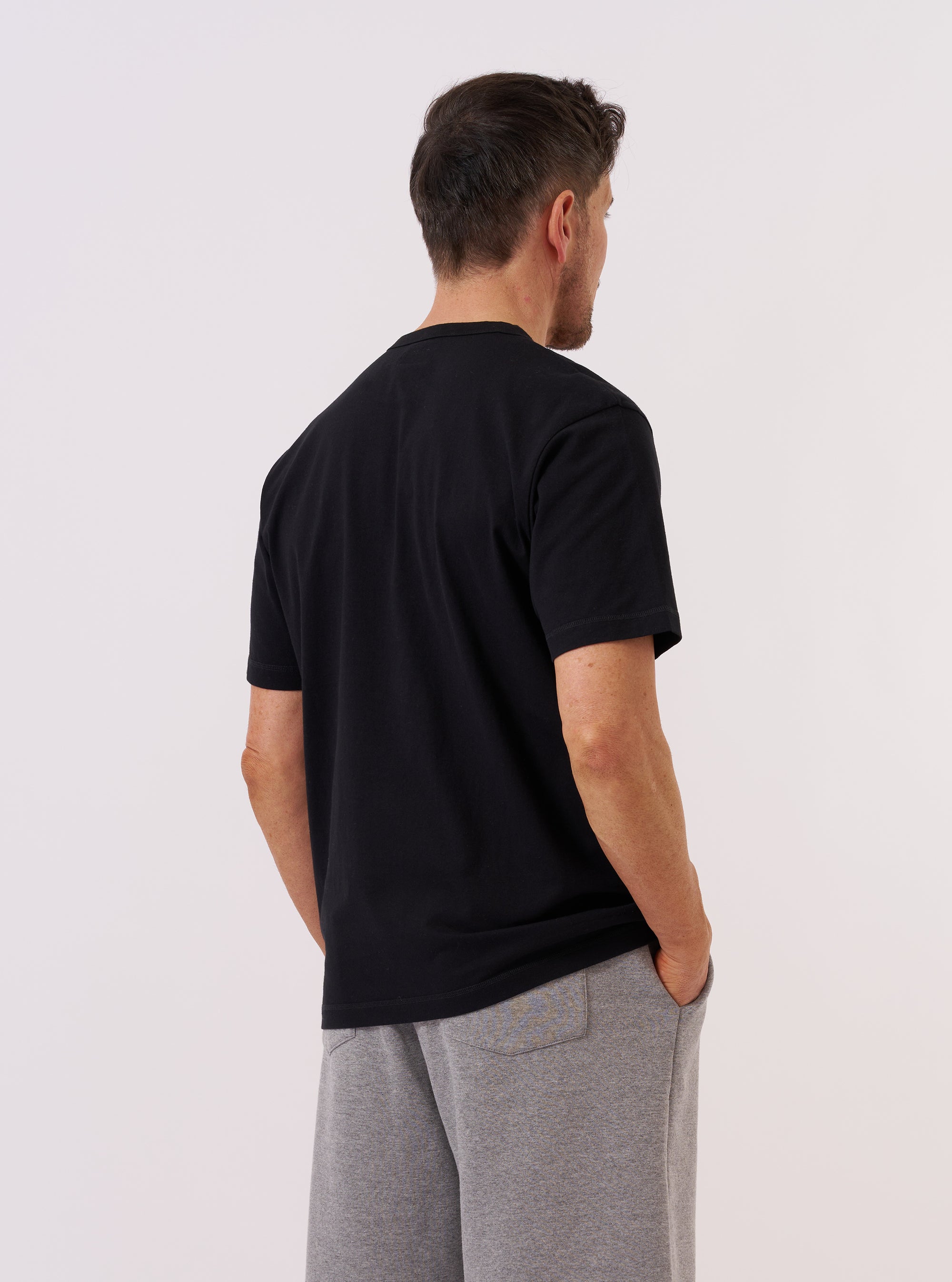 A model wears the Black Recycled/Bio Jersey Core Pocket Tee, shown from a side angle against a plain white background. The t-shirt features a classic crew neck, short sleeves, and a patch pocket on the left chest. The black jersey fabric has a smooth, soft texture and a relaxed fit.