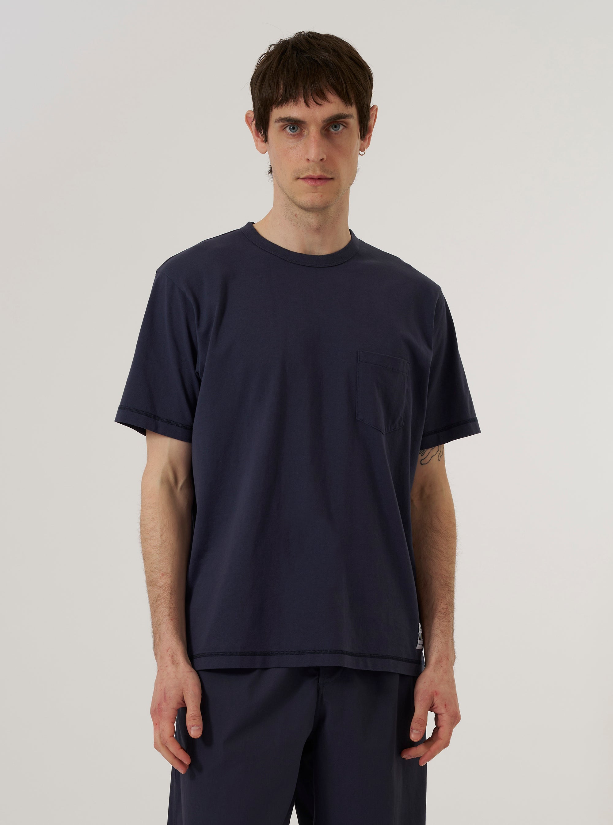 A model wears the Navy Recycled/Bio Jersey Core Pocket Tee against a plain, neutral background. The dark navy short-sleeved t-shirt features a crew neckline and a single patch pocket on the left chest. The image is a medium shot, showing the model from the waist up.
