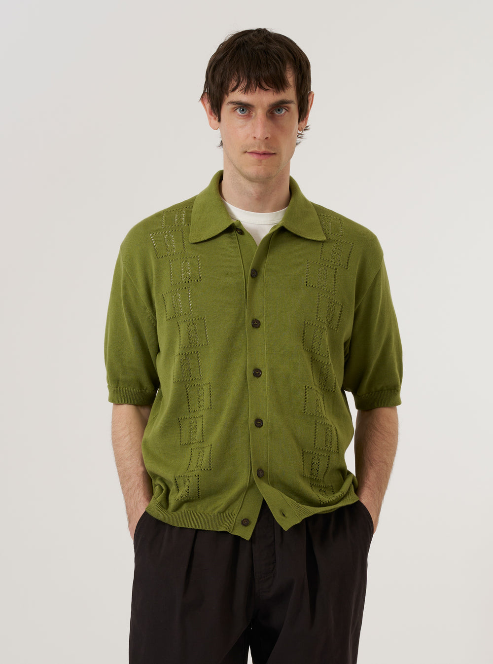 A model wears the Olive Cotton Lace Knit Jonny Knit Shirt against a plain white background. The shirt features a distinctive open-work lace knit pattern throughout, a classic collar, and a full-length button-down front. The olive green garment is worn over a light-colored t-shirt, highlighting the semi-sheer nature of the cotton lace fabric.