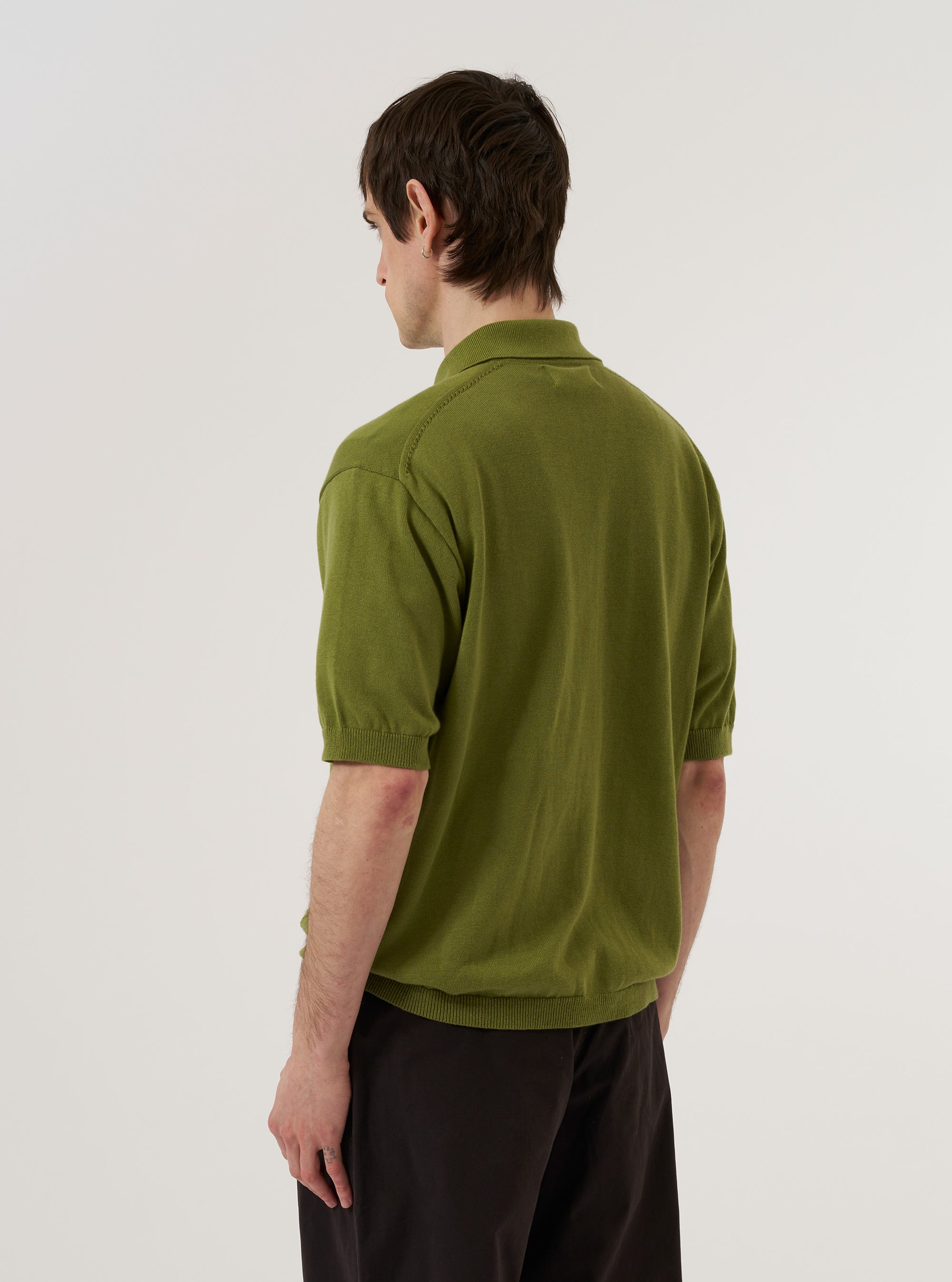 A model is wearing the Olive Cotton Lace Knit Jonny Knit Shirt, shown from the back against a plain white background. The shirt features a distinctive open lace-knit pattern throughout, a straight hem, and short sleeves. The model is wearing the olive green shirt over a light-colored top. No mannequin is present in the image.