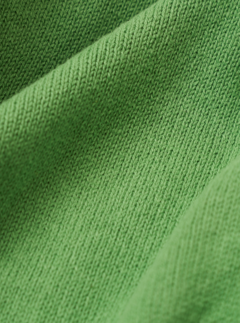 A close-up fabric swatch of the Green Eco Cotton Knit Pocket David Cardigan highlights the vibrant green color and the textured knit of the eco cotton material. The fabric fills the frame, providing a detailed view of the garment's weave and color.