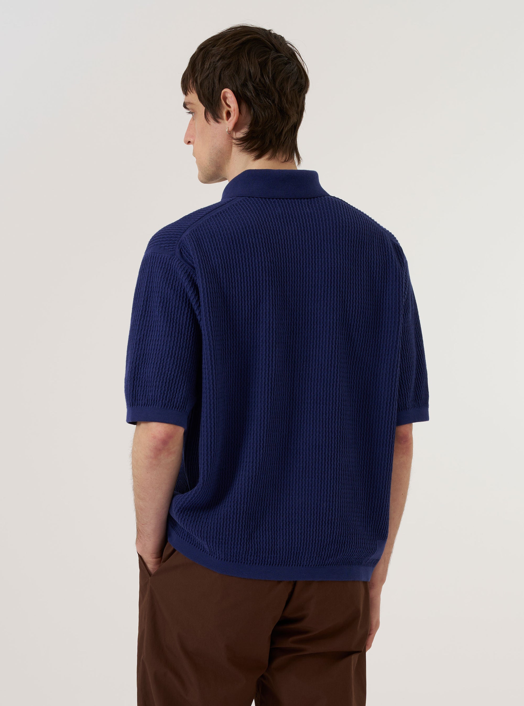 A model wears the Navy Fine Texture Tricot S/S Knit Polo, shown from a rear three-quarter angle against a plain, neutral background. The short-sleeved polo features a classic collar and a subtle, fine-texture tricot knit throughout the navy blue fabric.