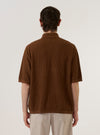 A model wears the Brown Fine Texture Tricot S/S Knit Polo against a plain white background. The image, captured from the chest up, highlights the polo's three-button placket, classic collar, and the intricate textured tricot knit of the brown cotton fabric. The model is wearing the polo buttoned up, showcasing its refined, structured fit. No mannequin is present in the image.