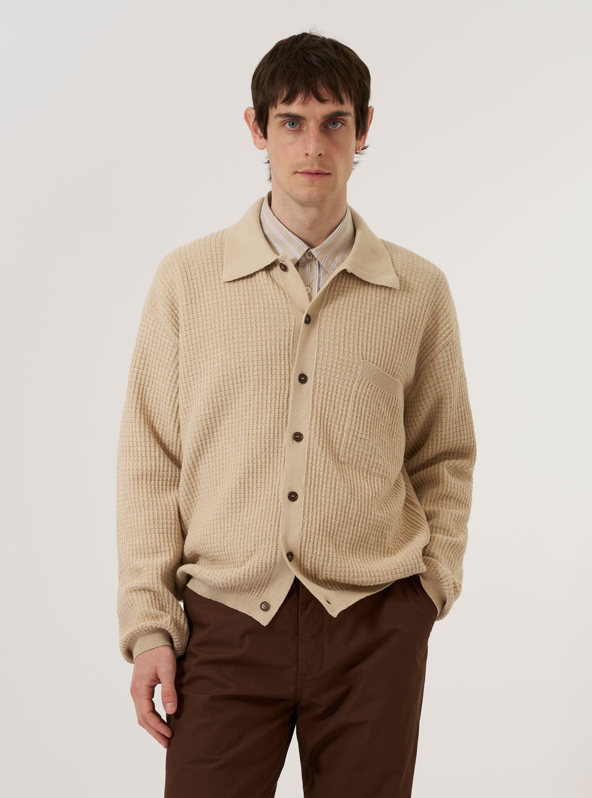 A model wears the Sand Eco Cotton Knit Jacket while standing against a plain white background. The jacket features a flat collar, a five-button front closure, and two large patch pockets at the waist. The knit fabric has a distinct, textured appearance in a neutral sand color.