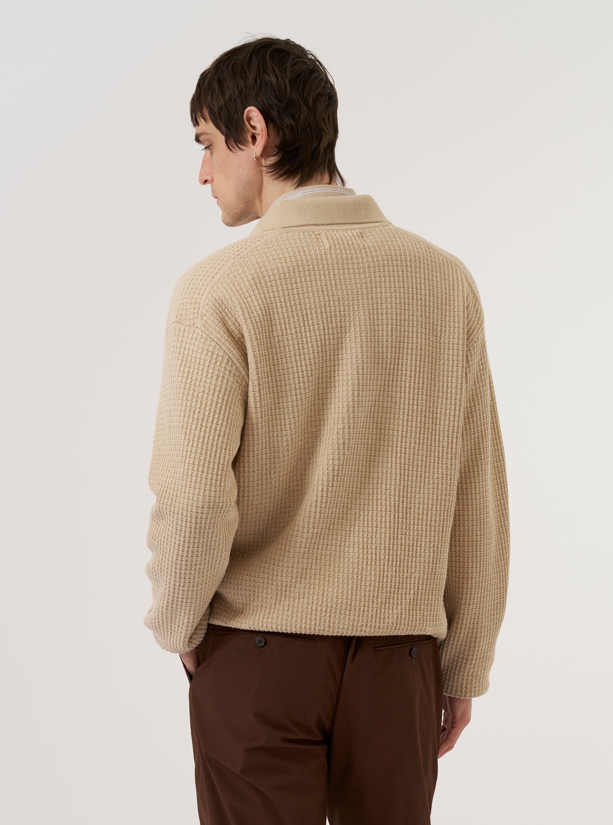 The Sand Eco Cotton Knit Jacket is worn by a model against a plain white background. The jacket is shown from a rear three-quarter angle, highlighting the textured knit fabric, a simple collar, and a straight hem. The model wears the jacket over a light-colored top and dark trousers.