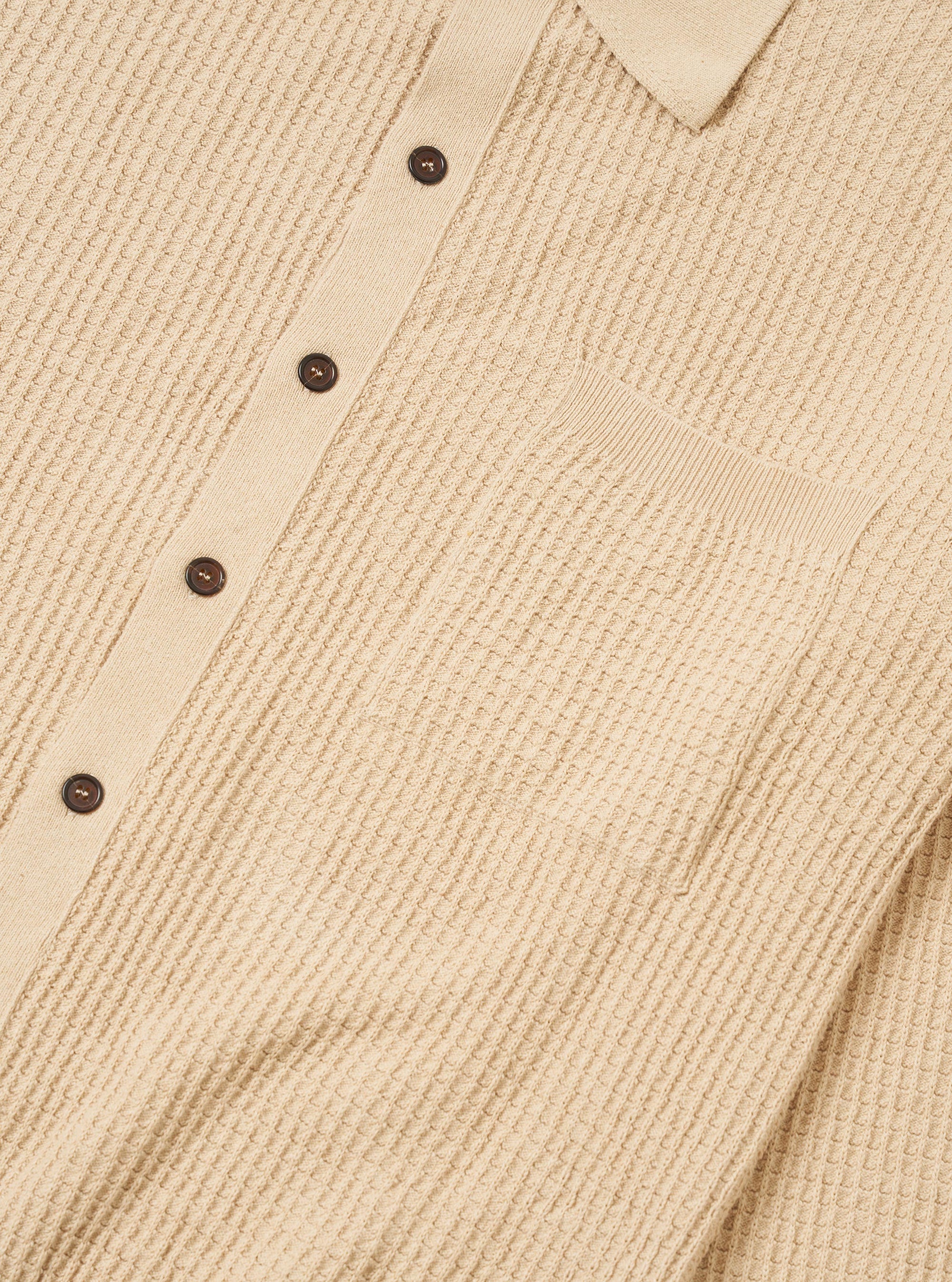 The Sand Eco Cotton Knit Jacket is shown from the back, worn by a model against a plain white background. The jacket features a clean, straight hem and a simple knit texture in a light sand color. The model is also wearing dark trousers, and the focus is on the fit and drape of the garment across the shoulders and back.