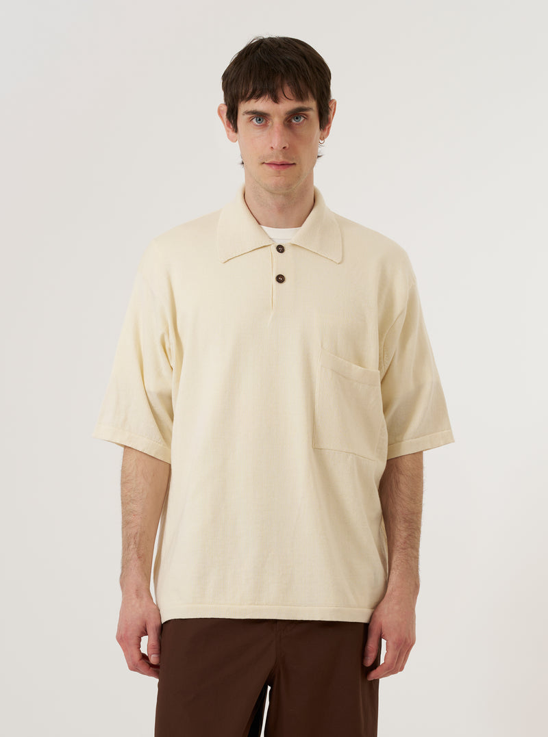 The Ecru Woolcot Bio S/S Pullover Knit Shirt II is worn by a model against a plain white background. The shirt features a classic collar, a three-button placket, and a single chest pocket. The short-sleeved garment is made from a textured ecru knit fabric and is worn tucked into dark trousers to highlight its relaxed fit.