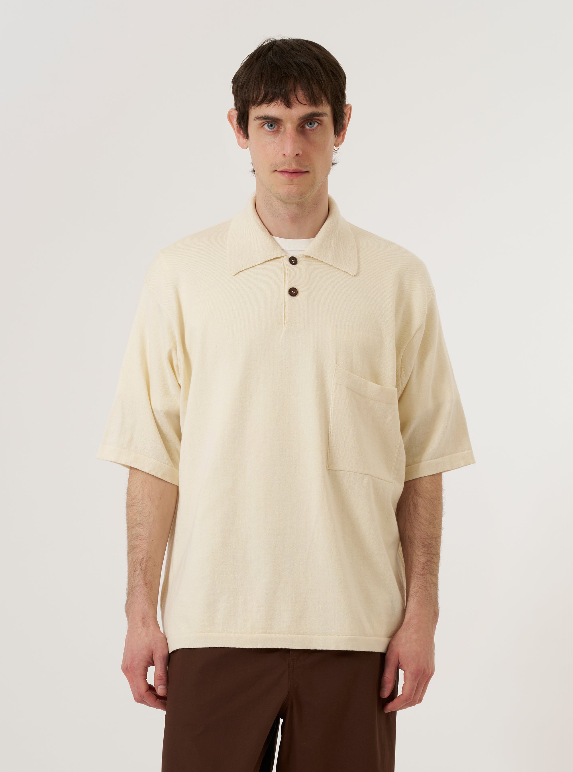 The Ecru Woolcot Bio S/S Pullover Knit Shirt II is worn by a model against a plain white background. The shirt features a classic collar, a three-button placket, and a single chest pocket. The short-sleeved garment is made from a textured ecru knit fabric and is worn tucked into dark trousers to highlight its relaxed fit.