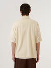 The Ecru Woolcot Bio S/S Pullover Knit Shirt II is worn by a model against a plain white background. The image, captured from the back, shows the model standing with hands in pockets, highlighting the garment's relaxed, boxy fit and short sleeves. The ecru-colored knit fabric appears soft and smooth, featuring a clean, straight hem. No mannequin is present in the image.
