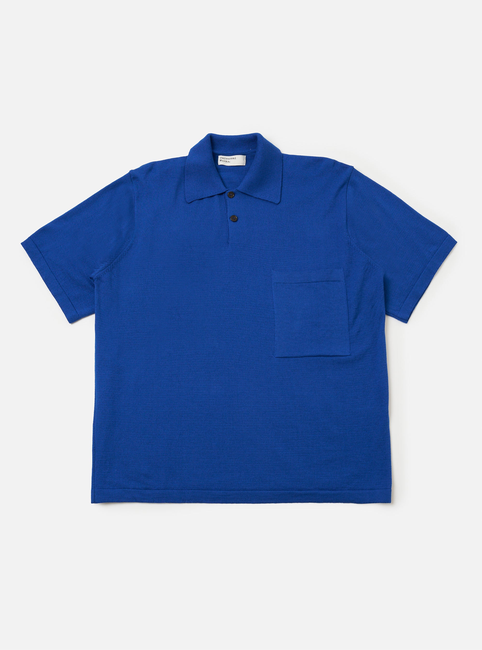 The Blue Woolcot Bio S/S Pullover Knit Shirt II is shown as a flat lay against a white background. The shirt features a boxy fit, short sleeves, a button placket, and a single chest pocket on the left side. The fabric is a smooth, solid blue knit.