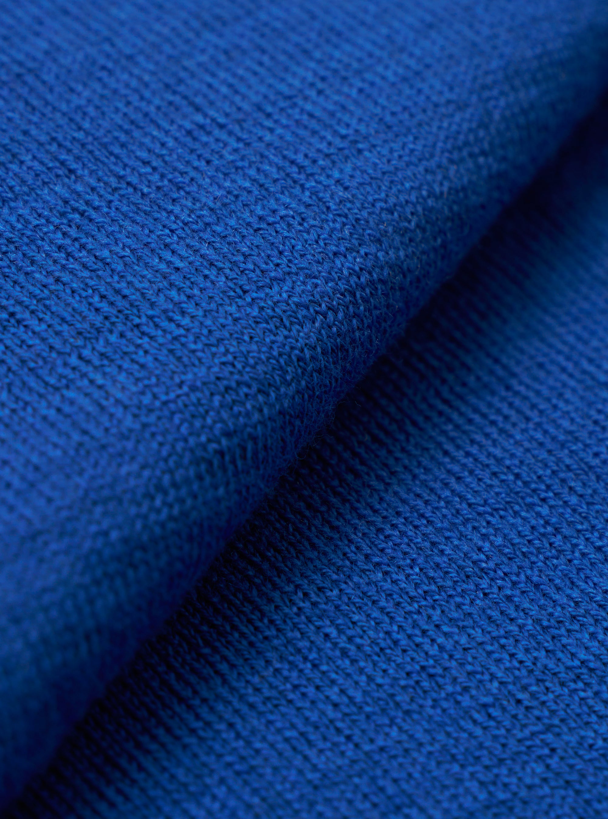 A close-up fabric swatch of the Blue Woolcot Bio S/S Pullover Knit Shirt II highlights the soft, fine-gauge texture of the blue knit material. The image focuses on the uniform weave of the merino and organic cotton blend.