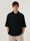 A model wears the Black Woolcot Bio S/S Pullover Knit Shirt II over a light-colored long-sleeved top against a plain white background. The shirt features a boxy, oversized fit, short sleeves, a classic collar, and a button placket. A single patch pocket is visible on the chest. The garment is constructed from a smooth, black merino and organic cotton blend knit.