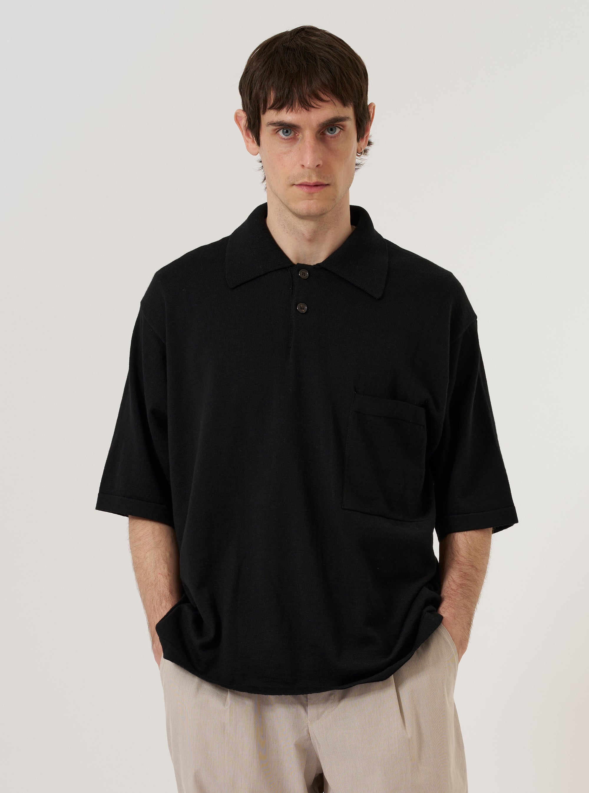 A model wears the Black Woolcot Bio S/S Pullover Knit Shirt II over a light-colored long-sleeved top against a plain white background. The shirt features a boxy, oversized fit, short sleeves, a classic collar, and a button placket. A single patch pocket is visible on the chest. The garment is constructed from a smooth, black merino and organic cotton blend knit.