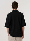 A model wears the Black Woolcot Bio S/S Pullover Knit Shirt II against a plain white background. The image, captured from the back at a slight angle, highlights the shirt's boxy fit, short sleeves, and smooth knit texture. The model is wearing the black pullover with light-colored trousers, showcasing the garment's relaxed silhouette and straight hem.