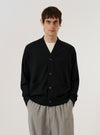 A model wears the Black Woolcot Bio Loose Cardigan II against a plain, neutral studio background. The cardigan features a relaxed, loose fit, a V-neckline, and a central button-front closure. The black knit fabric, made from a merino and organic cotton blend, has a soft and smooth appearance. The model is shown from the front, highlighting the garment's drape and classic knitwear construction.