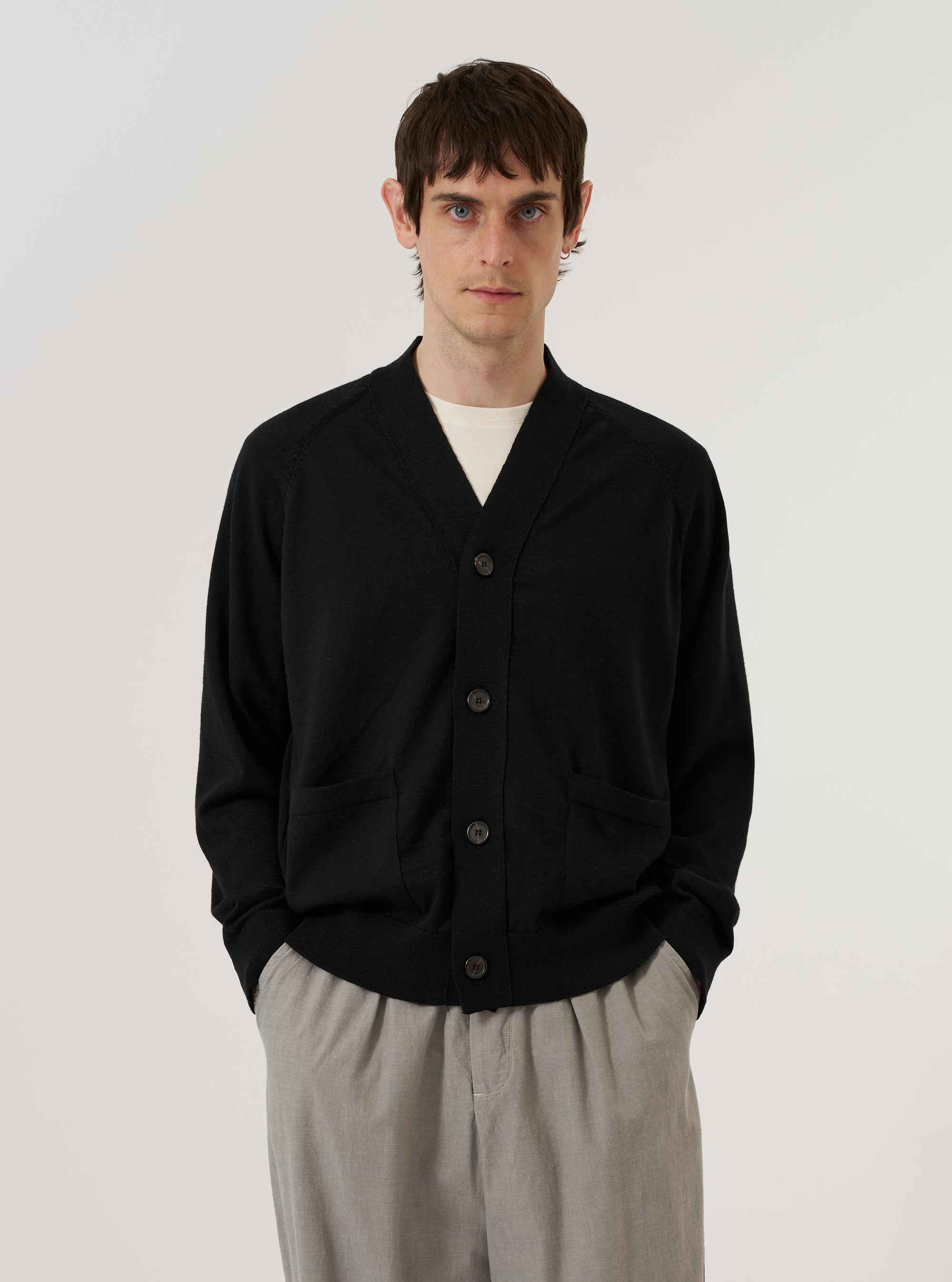 A model wears the Black Woolcot Bio Loose Cardigan II against a plain, neutral studio background. The cardigan features a relaxed, loose fit, a V-neckline, and a central button-front closure. The black knit fabric, made from a merino and organic cotton blend, has a soft and smooth appearance. The model is shown from the front, highlighting the garment's drape and classic knitwear construction.