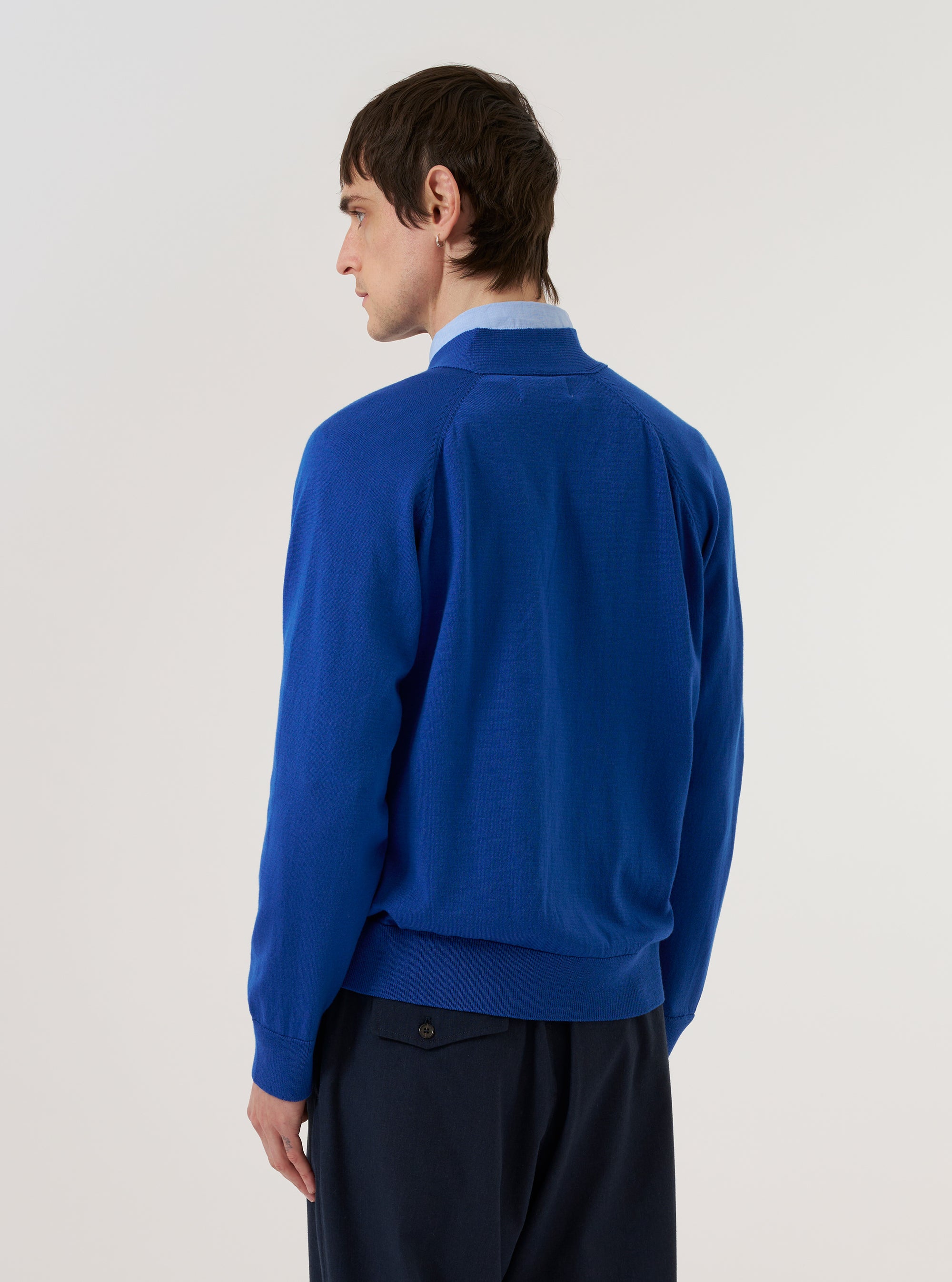A model wears the Blue Woolcot Bio Loose Cardigan II against a plain white background. The image, captured from the rear, highlights the garment's relaxed silhouette, V-neck collar, and fine gauge knit construction. The cardigan features ribbed detailing at the hem and cuffs, and is worn over a light-colored shirt.