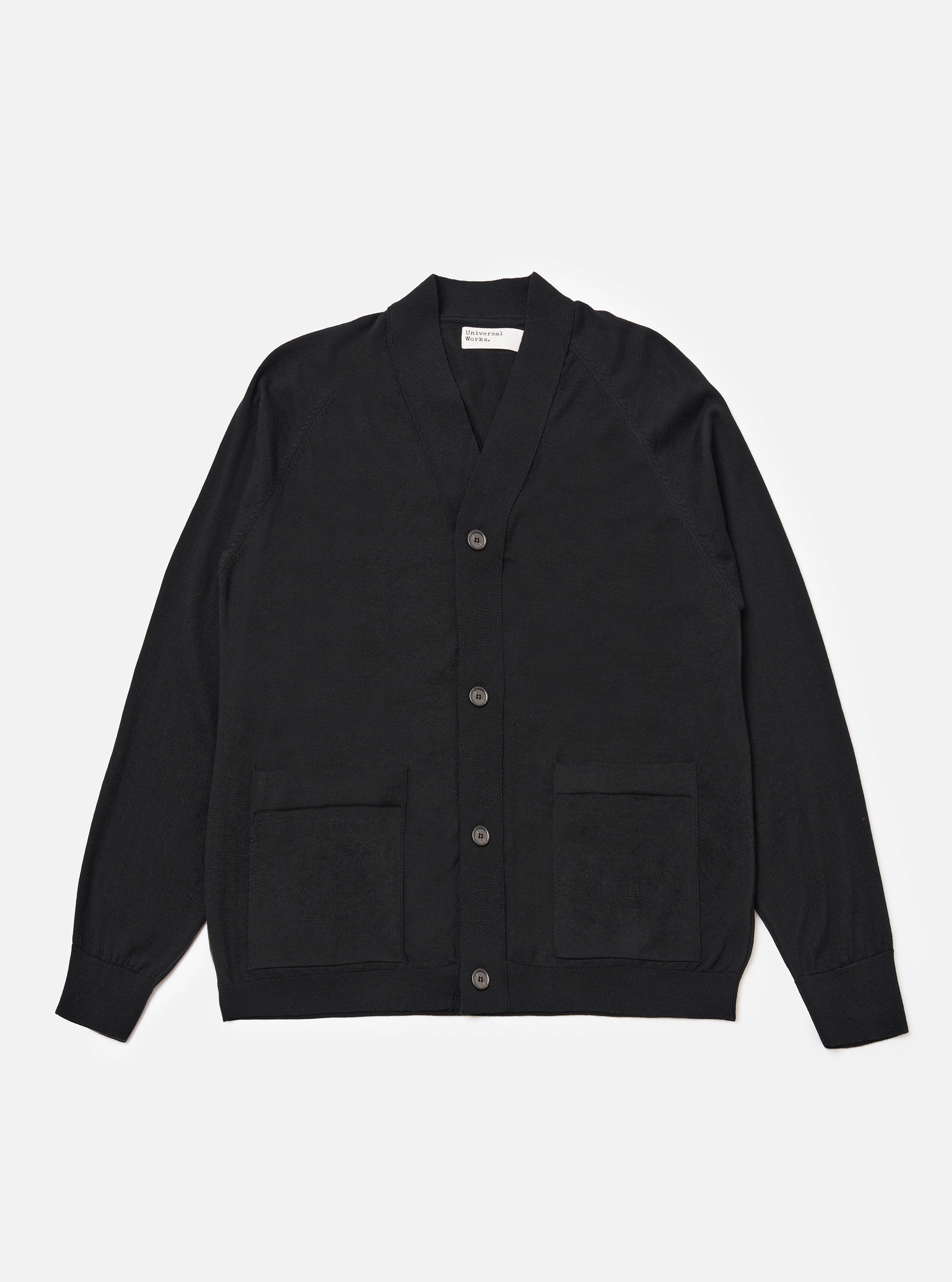 The Black Woolcot Bio Loose Cardigan II is shown as a flat lay against a plain white background. The cardigan features a V-neckline, a wide front button placket, and two patch pockets at the waist. The garment is constructed from a fine-gauge black knit and includes ribbed detailing on the cuffs and hem.